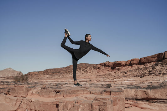 A woman wearing modest workout clothing performs a standing yoga pose on rocky terrain.