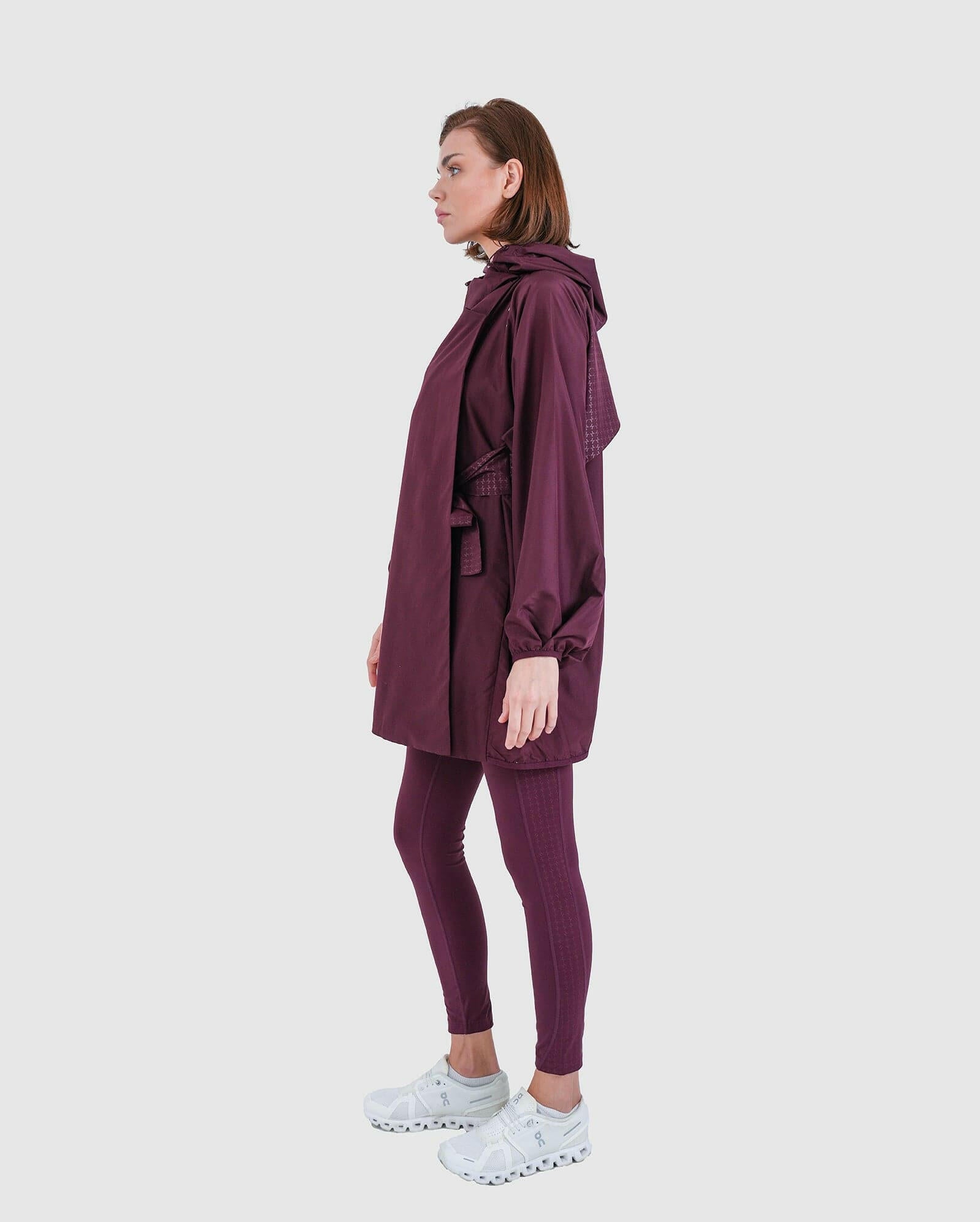 Side view of a woman wearing a maroon oversized cooldown coat by Qynda paired with matching leggings and white sneakers, posing against a neutral background.