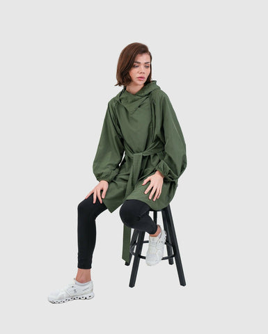 A model sitting on a black stool, wearing a Dark Olive cooldown coat by Qynda, black leggings, and white sneakers, posing with a contemplative expression against a neutral background.