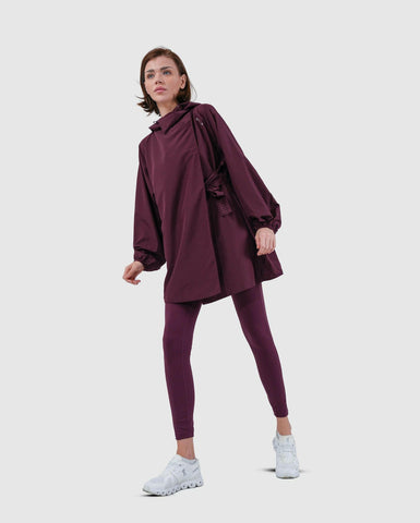 A woman twisted in a side wearing a maroon oversized cooldown coat by Qynda paired with matching leggings and white sneakers, posing against a neutral background.