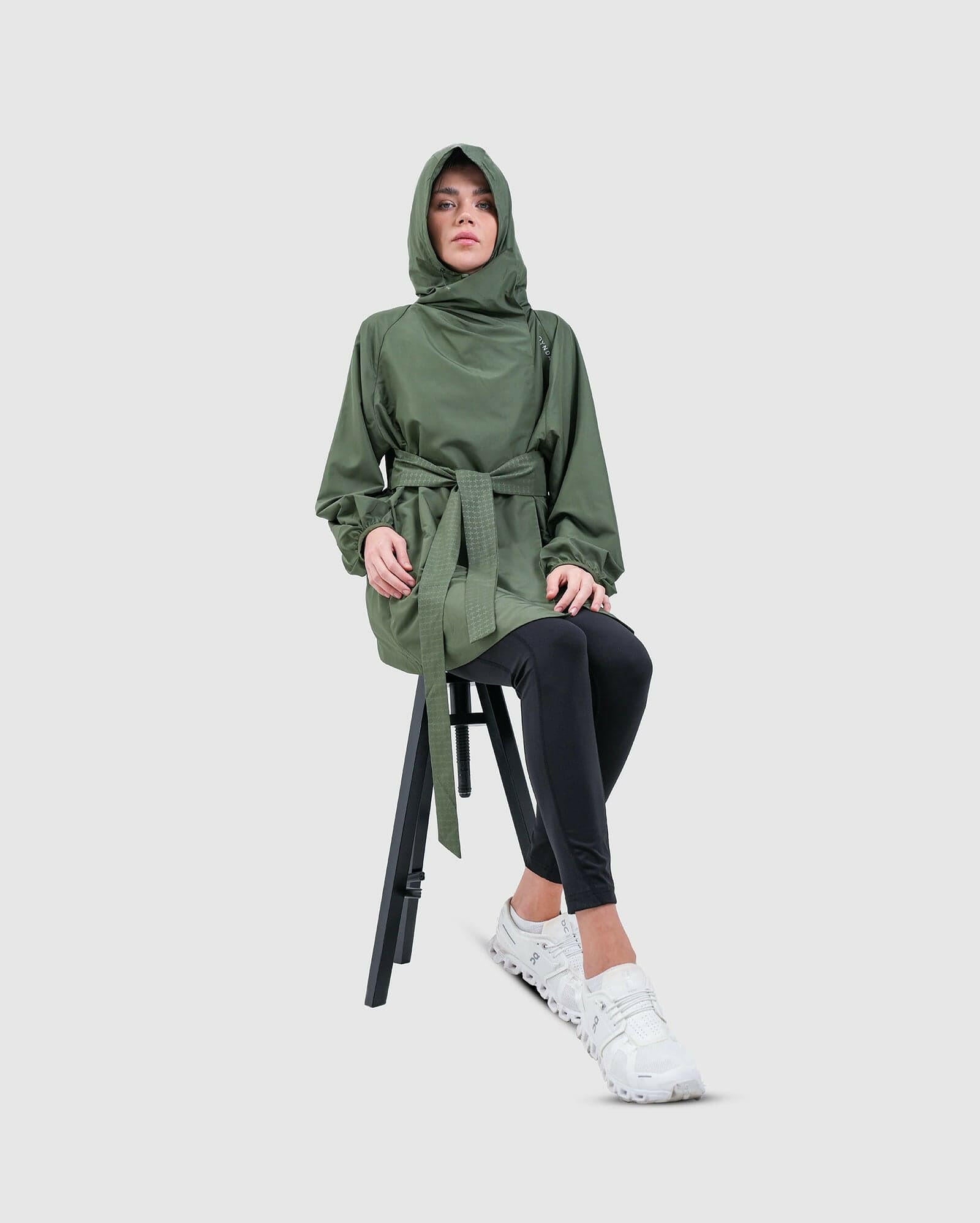 A model sitting on a black stool, wearing a Dark Olive hooded cooldown coat by Qynda, black leggings, and white sneakers, posing with a contemplative expression against a neutral background.