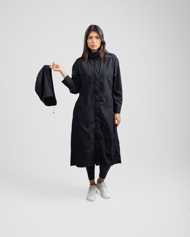 A model stands holding a black hood in one hand, wearing a modest black MAK LIGHT PARKA by Qynda made of lightweight fabric and white sneakers with breathable mesh. She is on a light grey background.