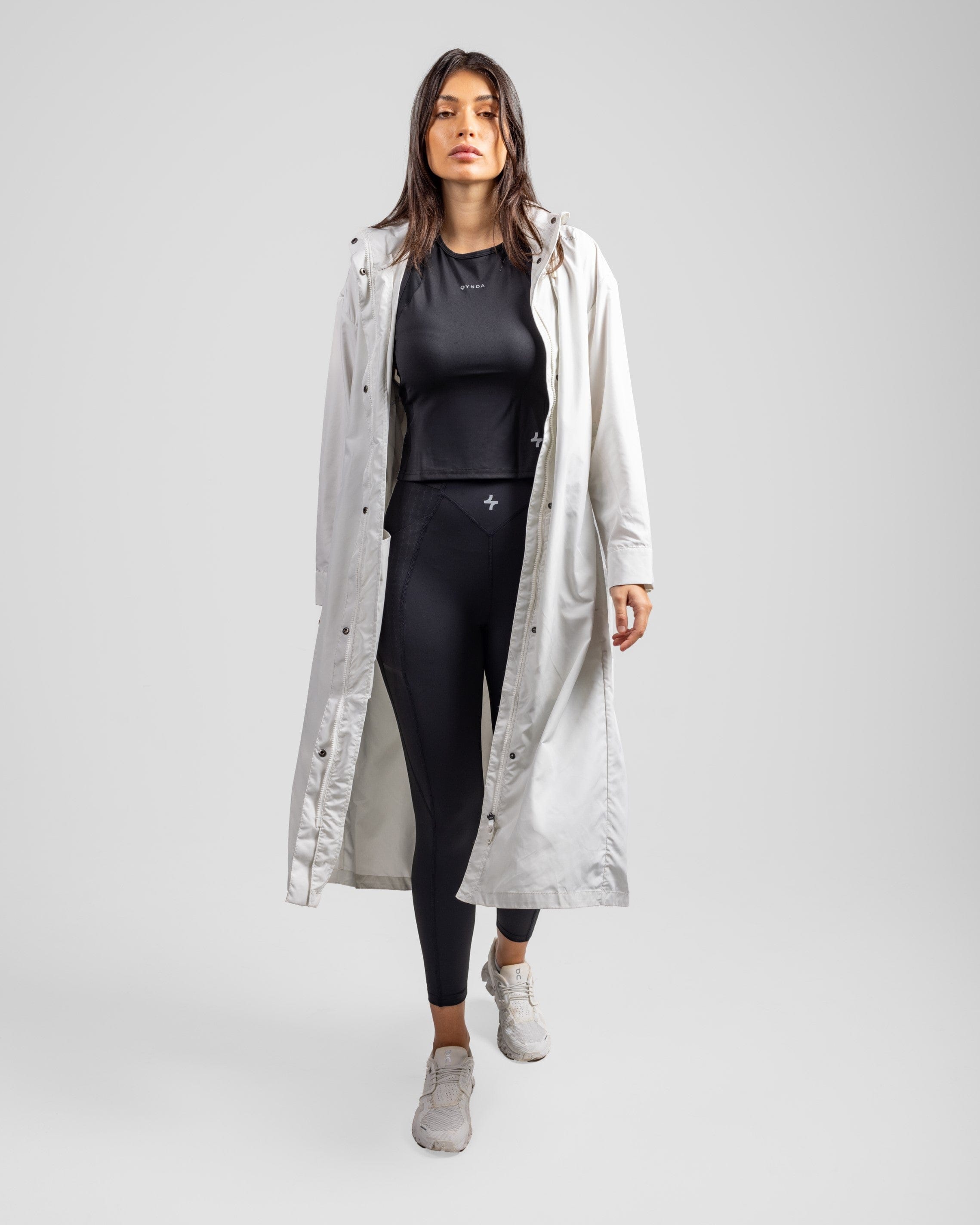A model, wearing a modest Off White MAK LIGHT PARKA by Qynda, made of lightweight fabric and gray sneakers, set against a plain light background.