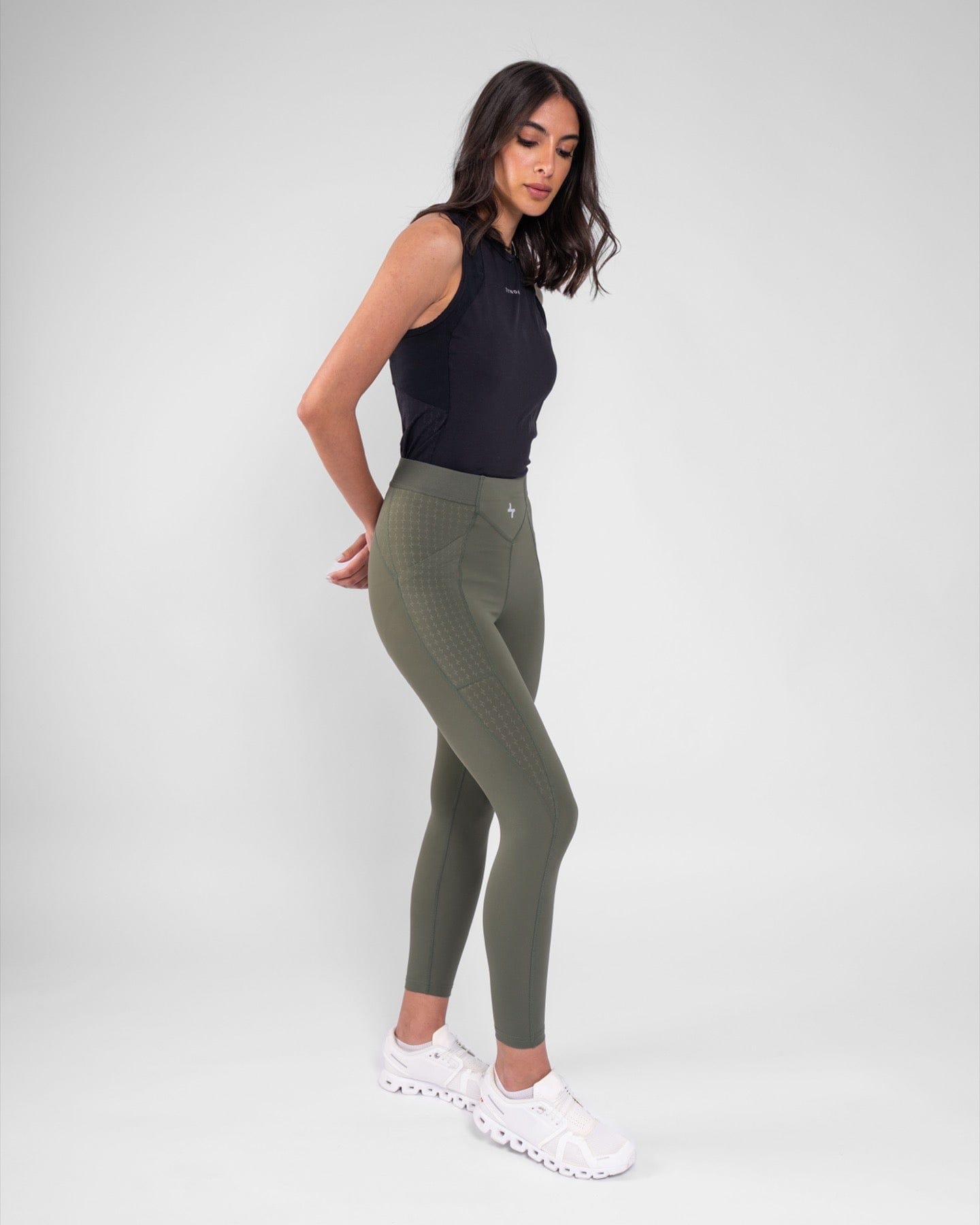 A woman seen posing, wearing a black tank top and olive THABYA LEGGINGS leggings by qynda, standing against a light grey background.