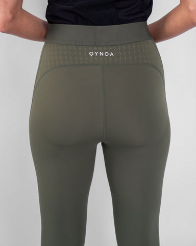 A woman standing with her back turned towards the camera, wearing a Olive modest THABYA LEGGINGS against a neutral gray background.