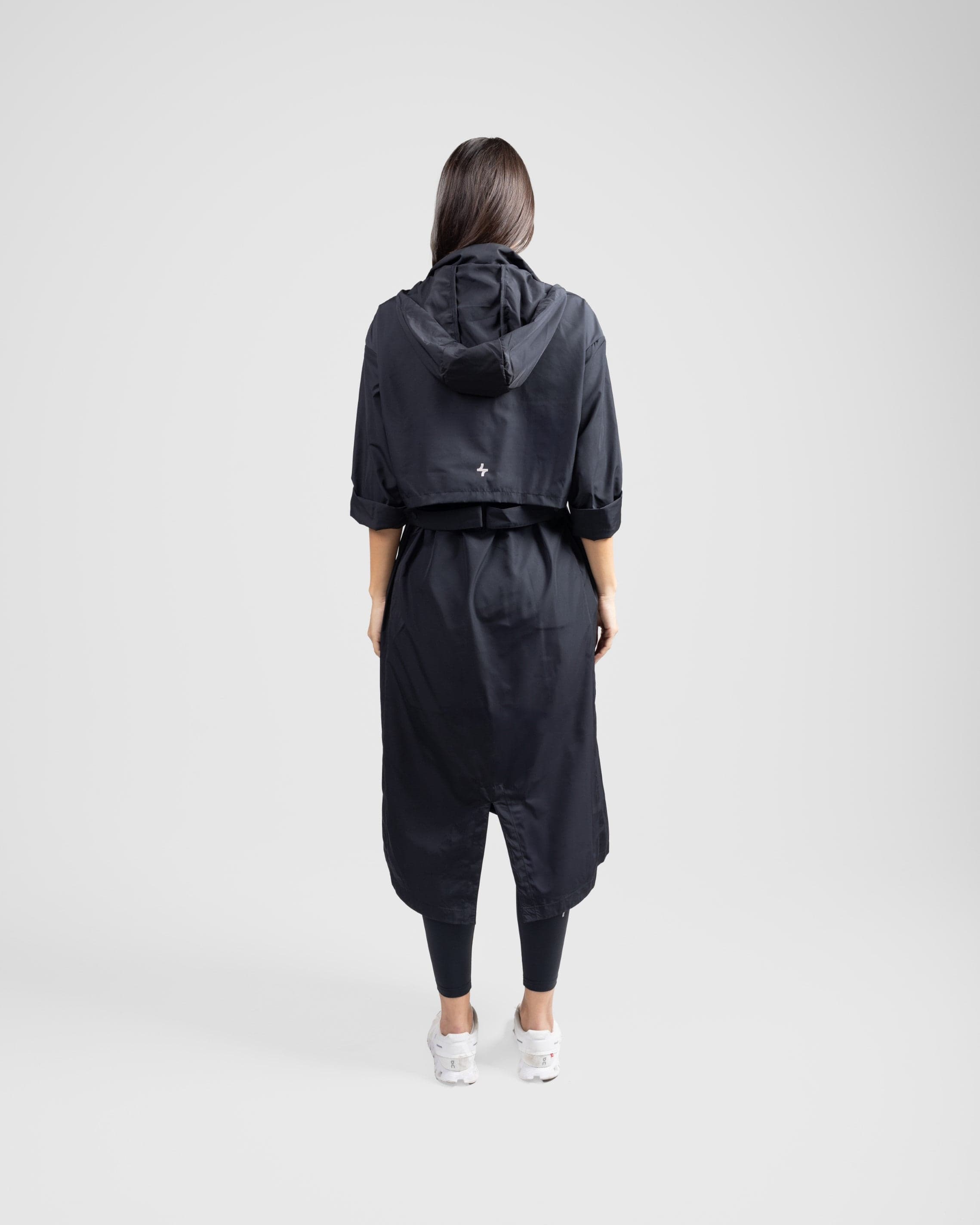 A model from behind wearing a modest black MAK LIGHT PARKA by Qynda with a hood and white breathable mesh sneakers, walking to the right on a plain light gray background.