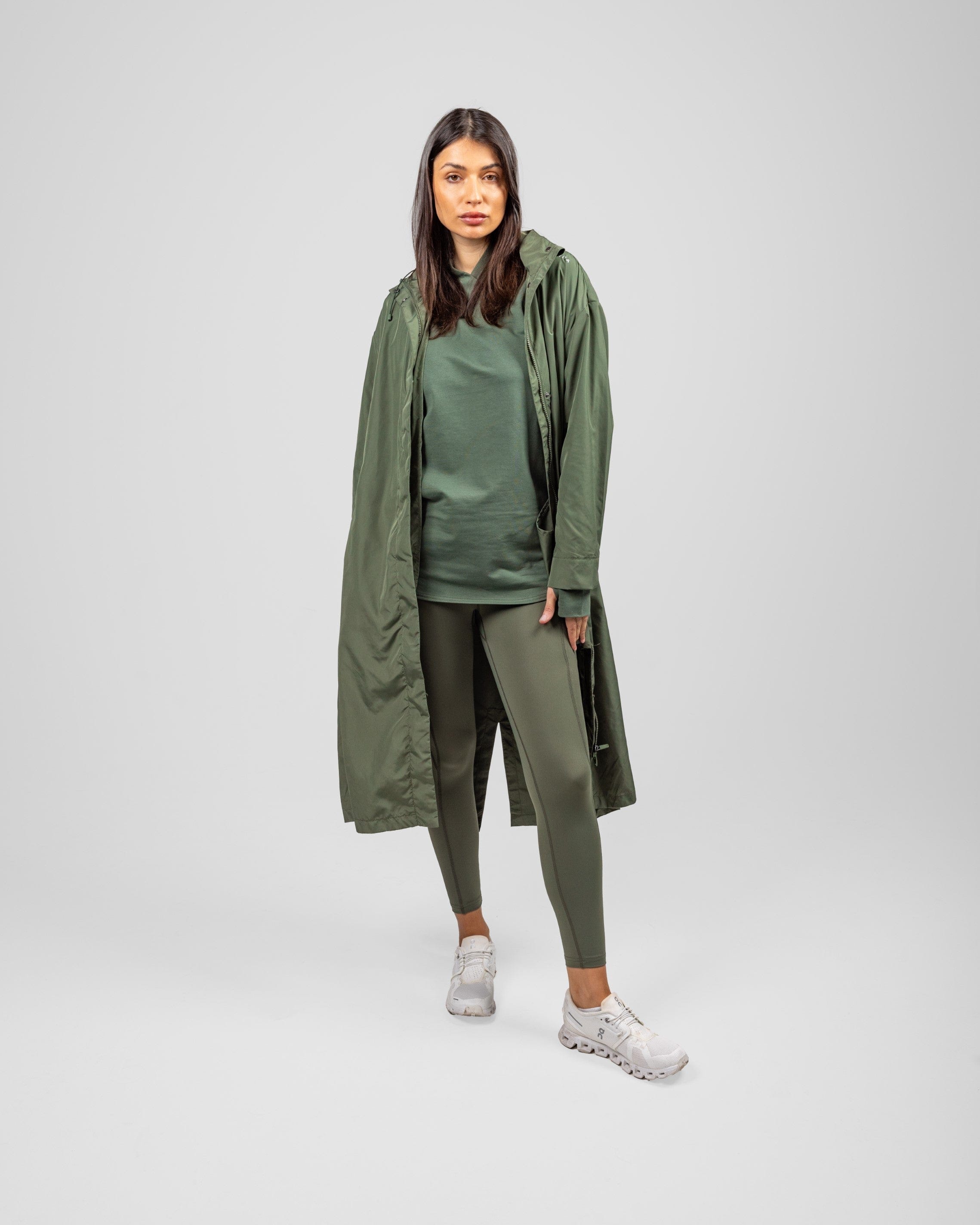 A model in an Olive MAK LIGHT PARKA by Qynda made of lightweight fabric and leggings, posing, standing against a light gray background.