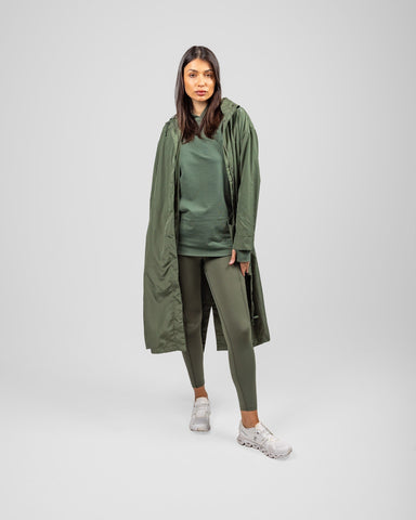 A model in an Olive MAK LIGHT PARKA by Qynda made of lightweight fabric and leggings, posing, standing against a light gray background.