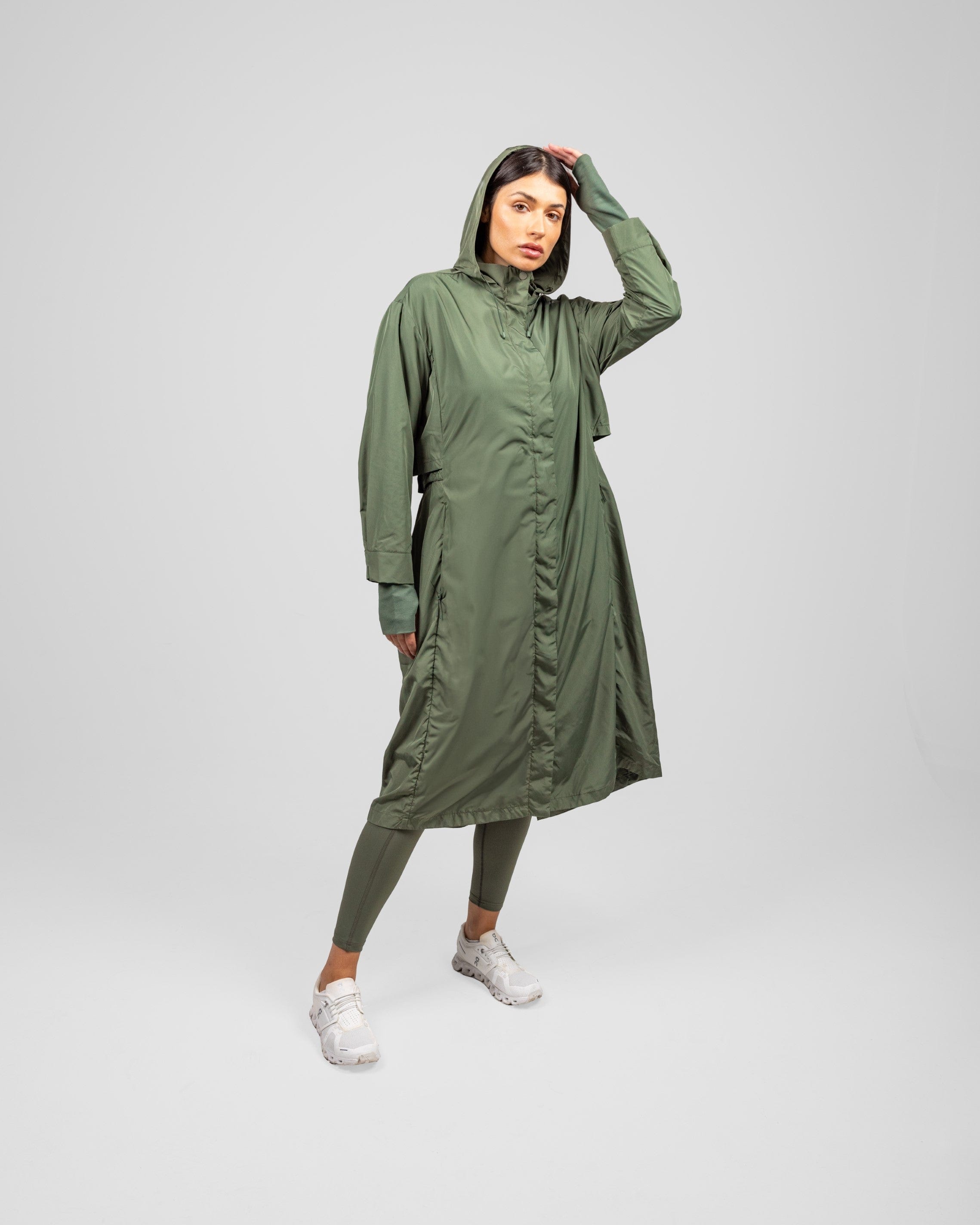 A model in an Olive MAK LIGHT PARKA by Qynda made of lightweight fabric and leggings, posing with one hand on her head, standing against a light gray background.