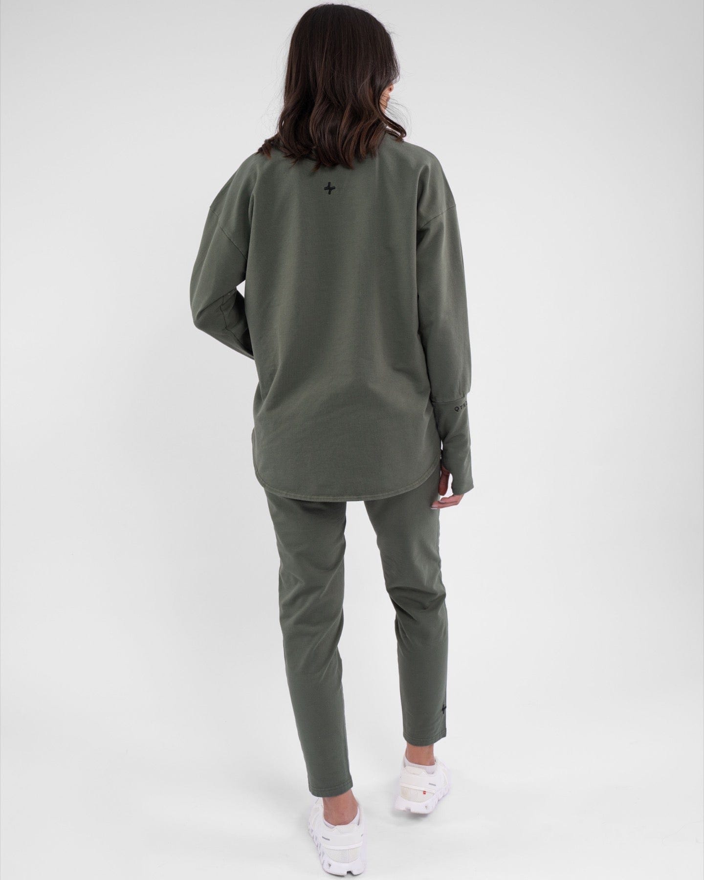 A woman stands with her back to the camera, showcasing a long-sleeve, Olive-colored REHAL SWEATER paired by qynda with matching leggings, both featuring cooling fabric technology. 