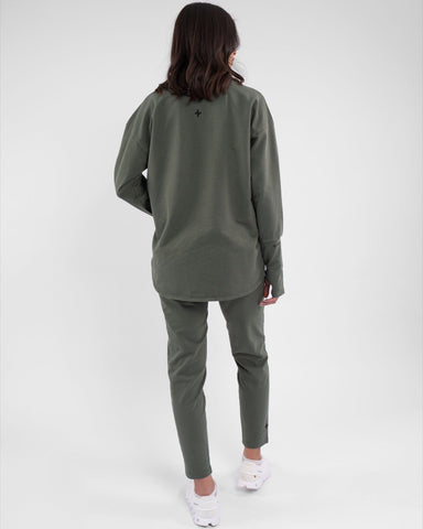 A woman stands with her back to the camera, showcasing a long-sleeve, Olive-colored REHAL SWEATER paired by qynda with matching leggings, both featuring cooling fabric technology. 