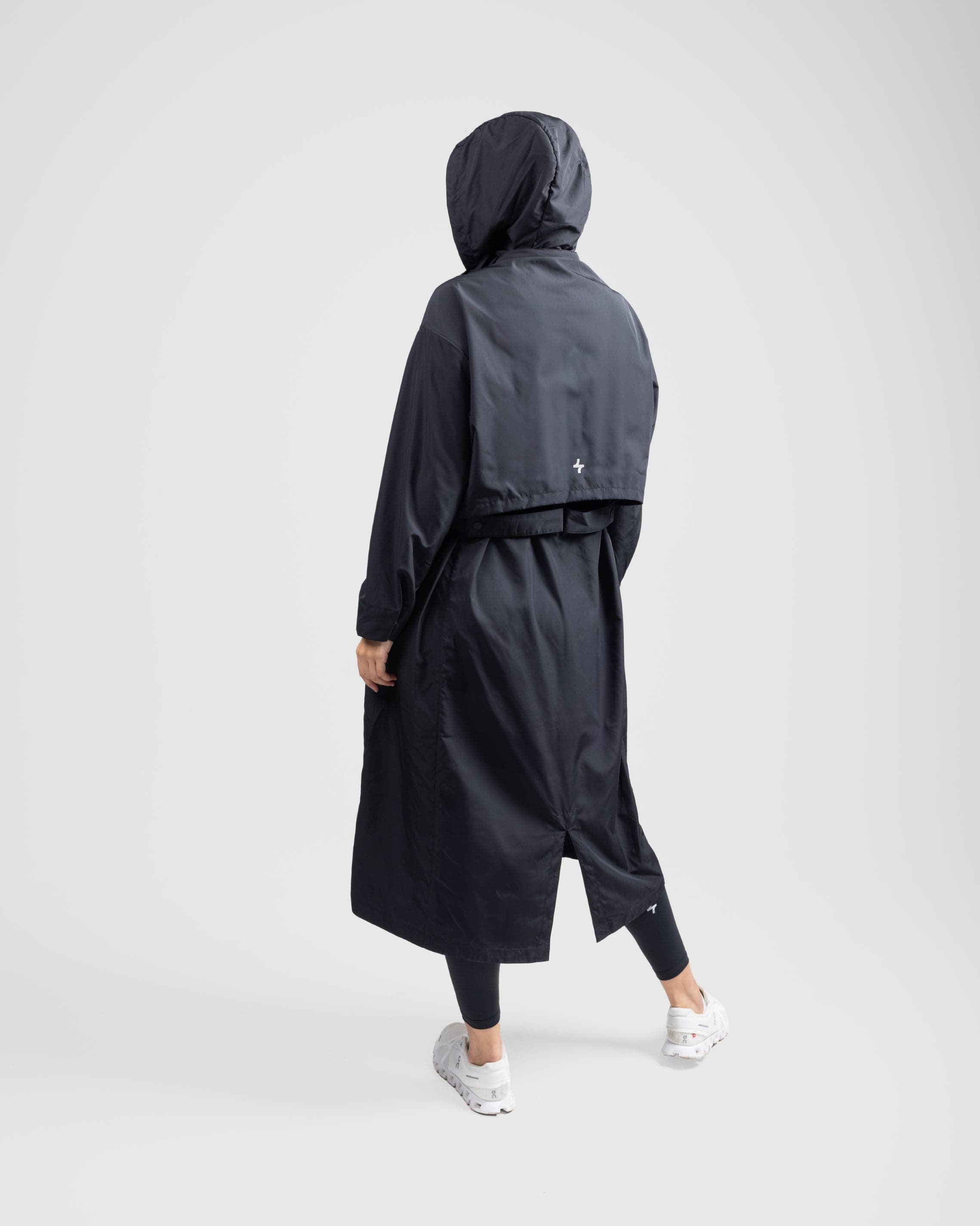 A model from behind wearing a black MAK LIGHT PARKA by Qynda with a hood and white breathable mesh sneakers, walking to the right on a plain light gray background.