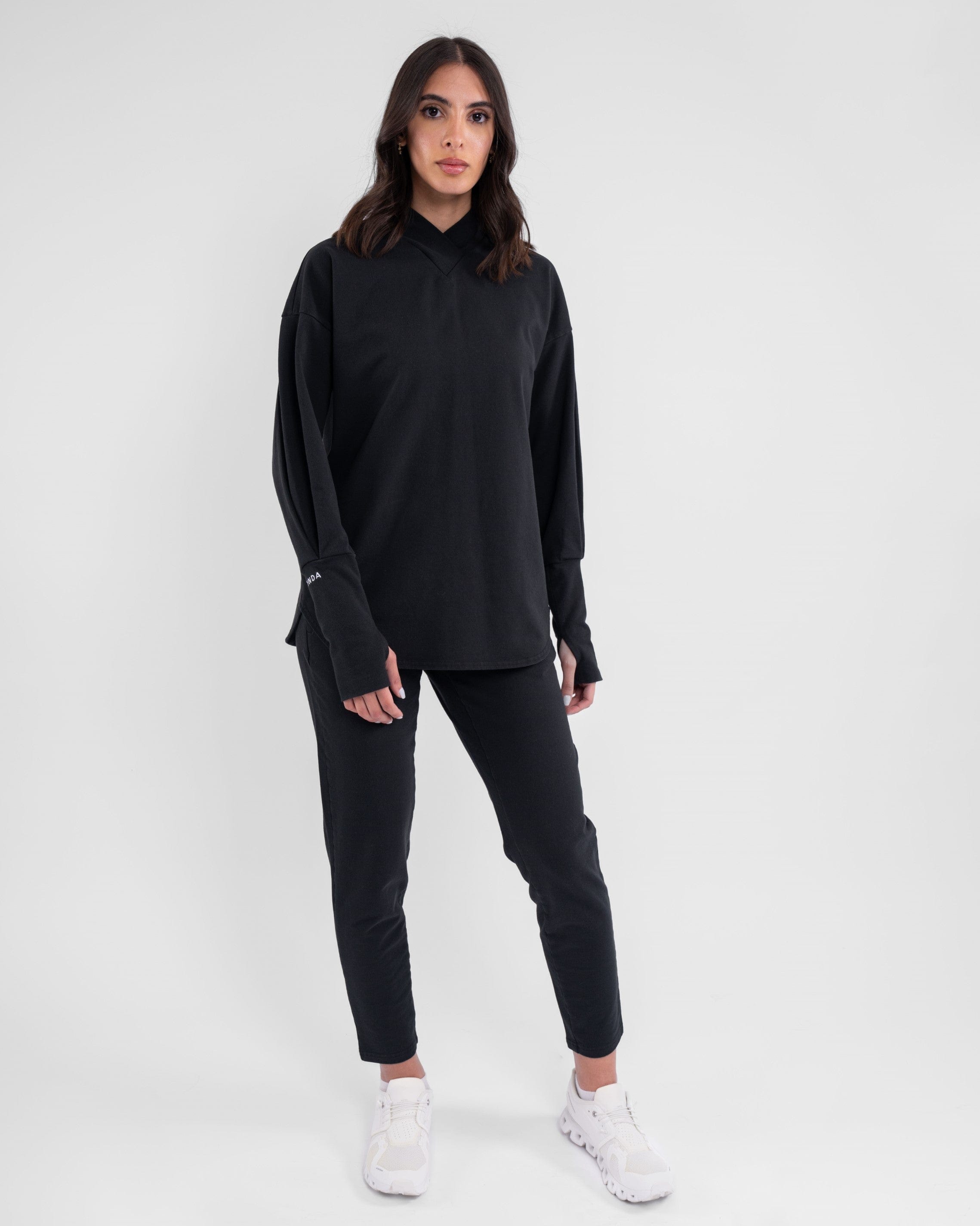 A woman stands confidently modeling a modest athletic CANTARA PANTS consisting of a long-sleeve shirt and slim pants, both made from moisture-wicking cotton terry loop fabric, complemented by a pair of white shoes.
