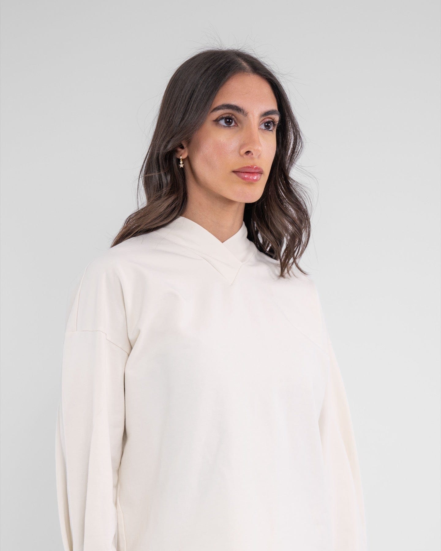 A woman with dark hair wearing an Off White modest SWEATER made of brrr° material, featuring moisture resistance and rapid drying capabilities, with the brand "qynda" on it, looking.