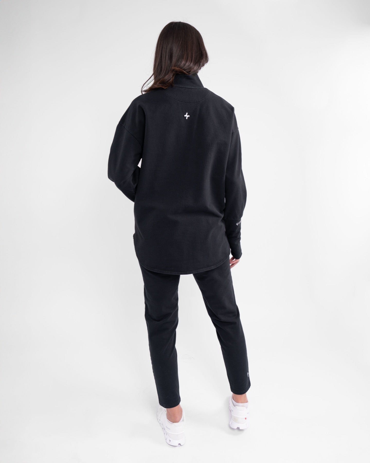 A woman stands with her back to the camera, showcasing a long-sleeve, black-colored modest athletic top paired with matching leggings, both featuring cooling fabric technology. 