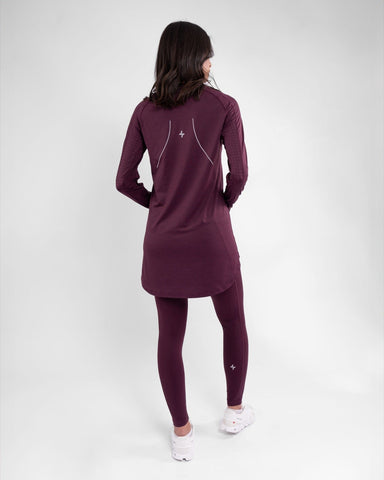 A woman stands with her back to the camera, showcasing a long-sleeve, plum-colored modest athletic top paired with matching leggings, both featuring cooling fabric technology. 