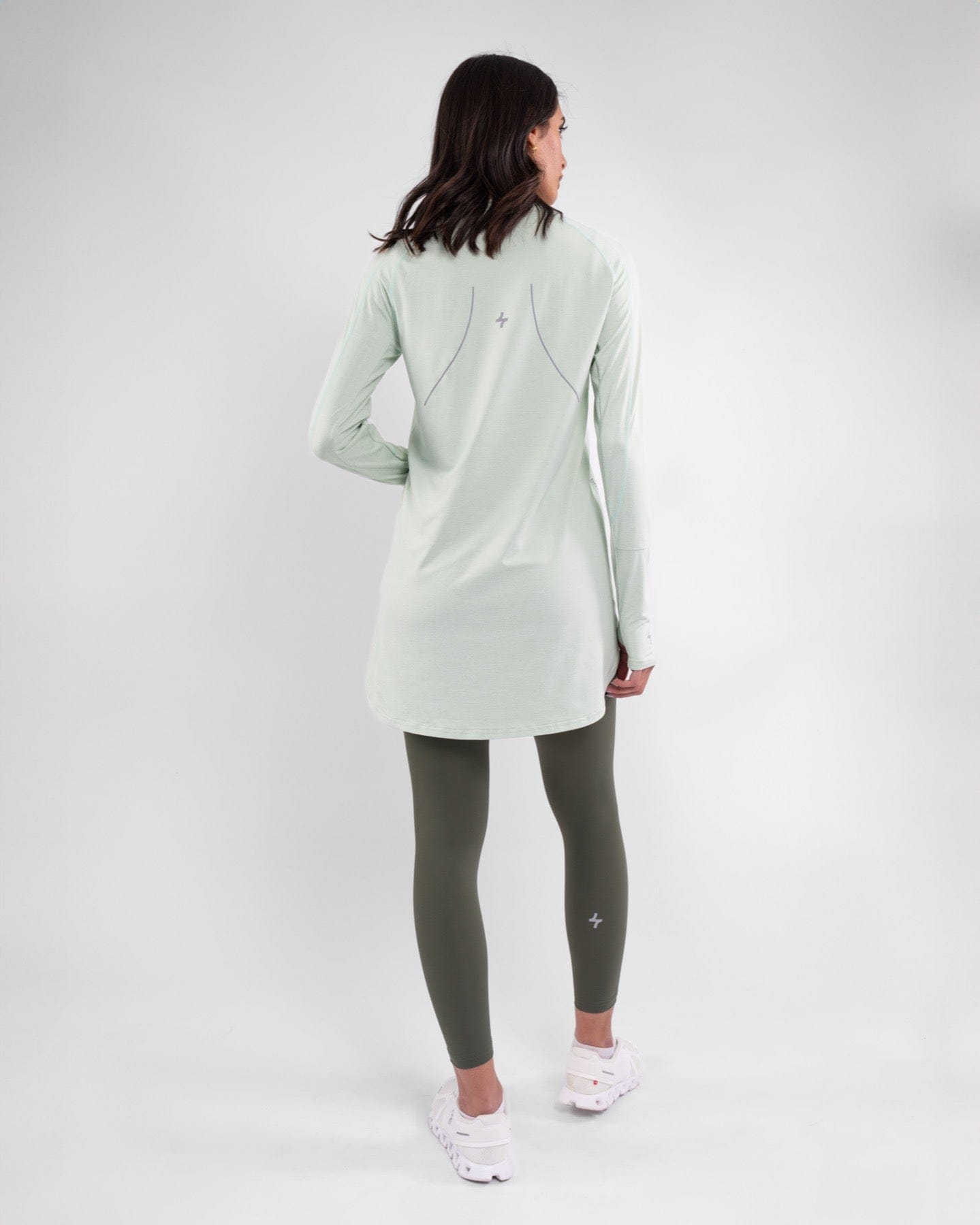 Woman posing in a light green, lightweight long-sleeve modest activewear top with cooling fabric technology and dark green, moisture-resistant leggings with a logo, paired with white sneakers, against a neutral background.