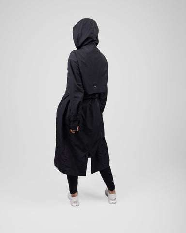 A model from behind wearing a black MAK LIGHT PARKA by Qynda with a hood and white breathable mesh sneakers, walking to the right on a plain light gray background.
