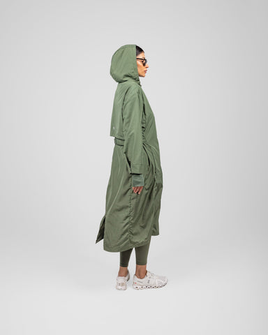 A model wearing an Olive MAK LIGHT PARKA by Qynda made of lightweight fabric and sunglasses stands in profile on a gray background, looking to the right. 