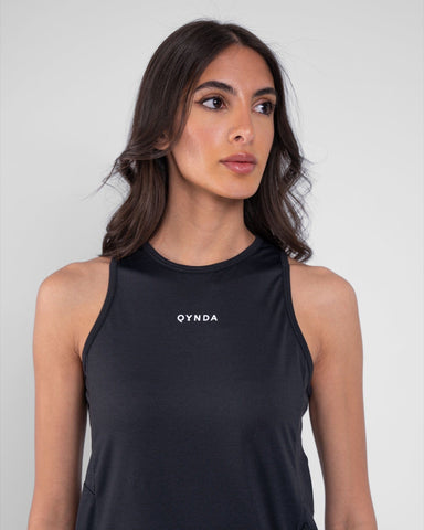 A woman with dark hair wearing a black sleeveless modest athletic top made of brrr° material, featuring moisture resistance and rapid drying capabilities, with the brand "qynda" on it, looking.