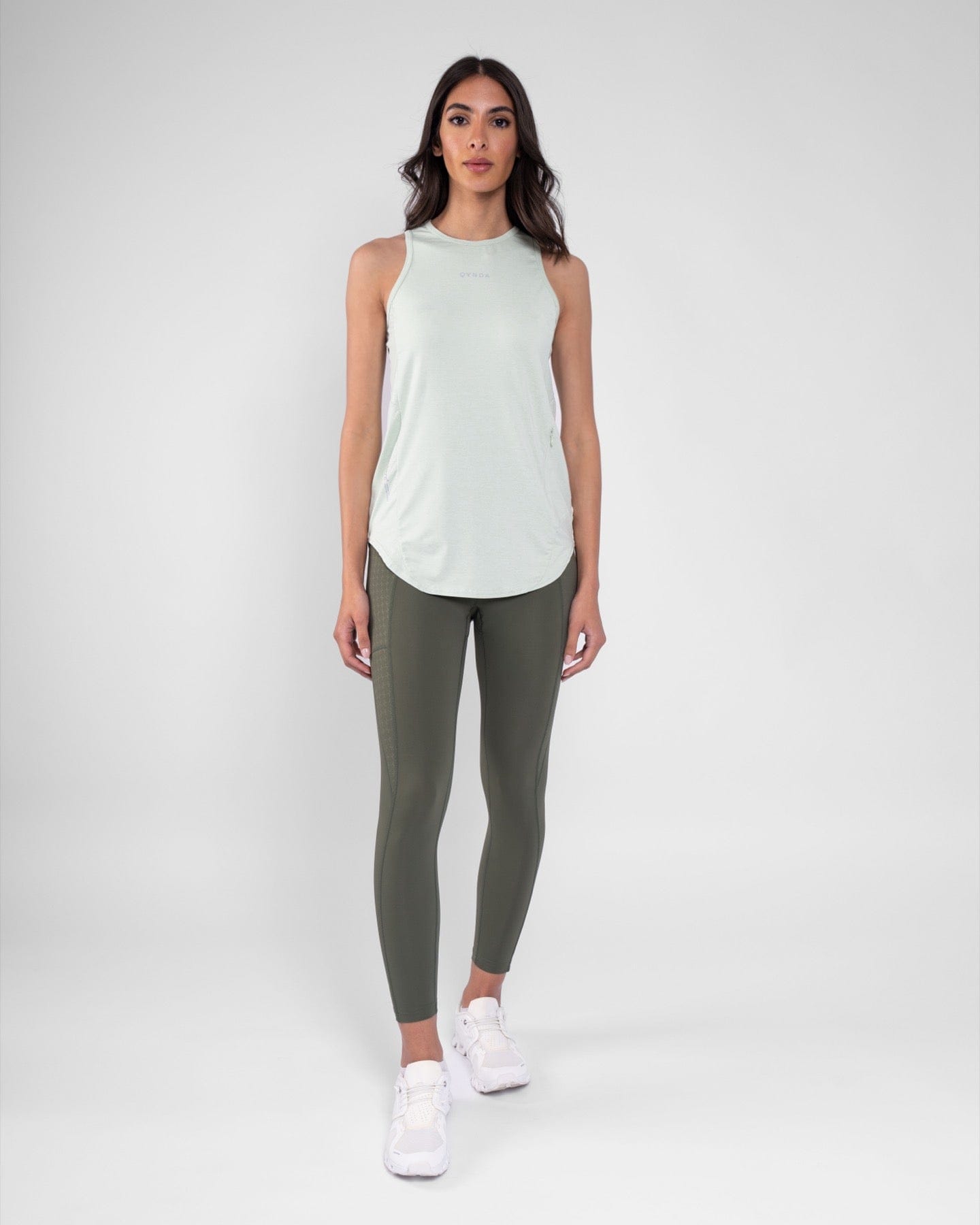 A woman is seen, wearing a RUH TANK TOP and Sage leggings by qynda, standing against a light grey background.