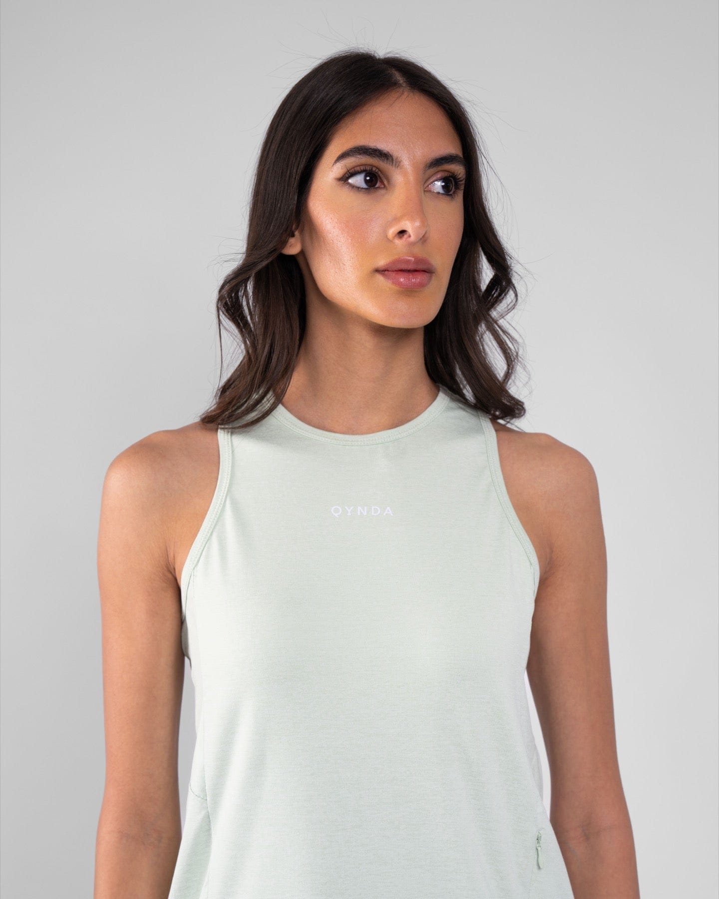 A woman with dark hair wearing a Sage sleeveless modest RUH TANK TOP made of brrr° material, featuring moisture resistance and rapid drying capabilities, with the brand "qynda" on it, looking.
