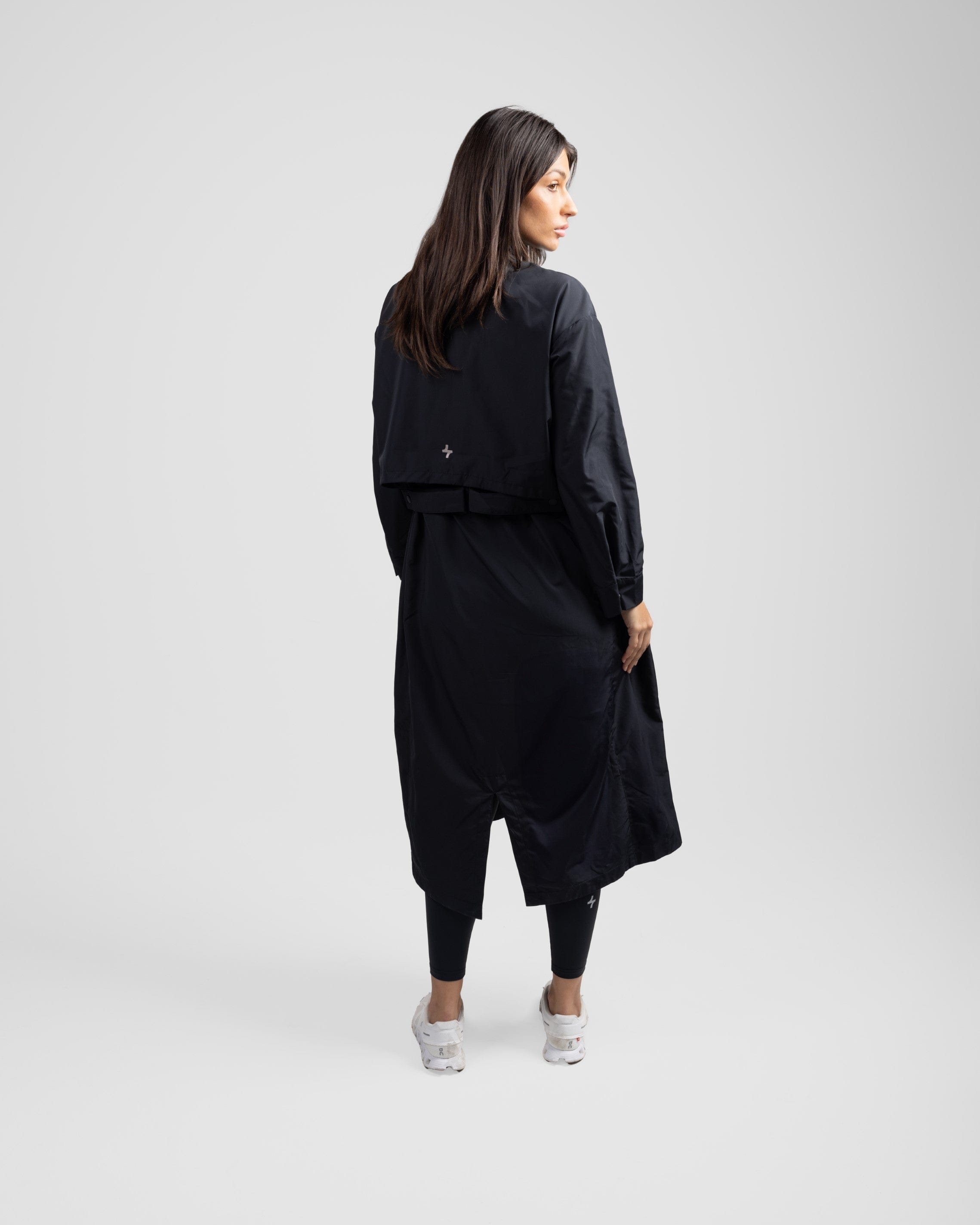 A model viewed from behind, turns her head to her left, wearing a stylish black modest MAK LIGHT PARKA by Qynda with a dual-direction zipper and sneakers, standing against a light gray background.