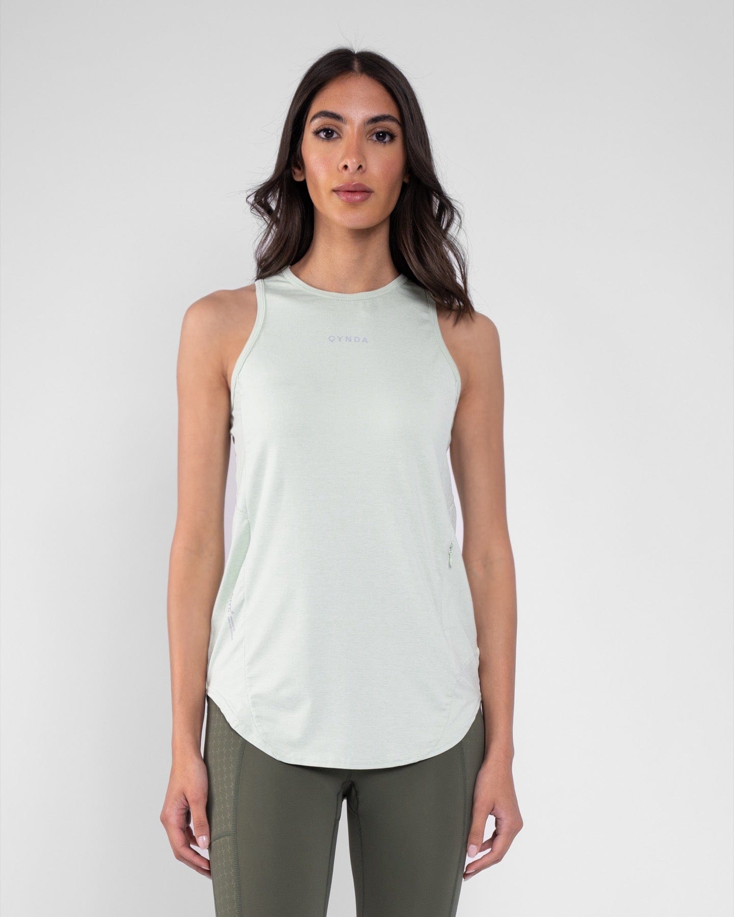 A woman in a light tank top and sage leggings made with brrr° material for body temperature regulation, paired with white sneakers against a clean, grey background, showcasing a casual modest athletic outfit by qynda