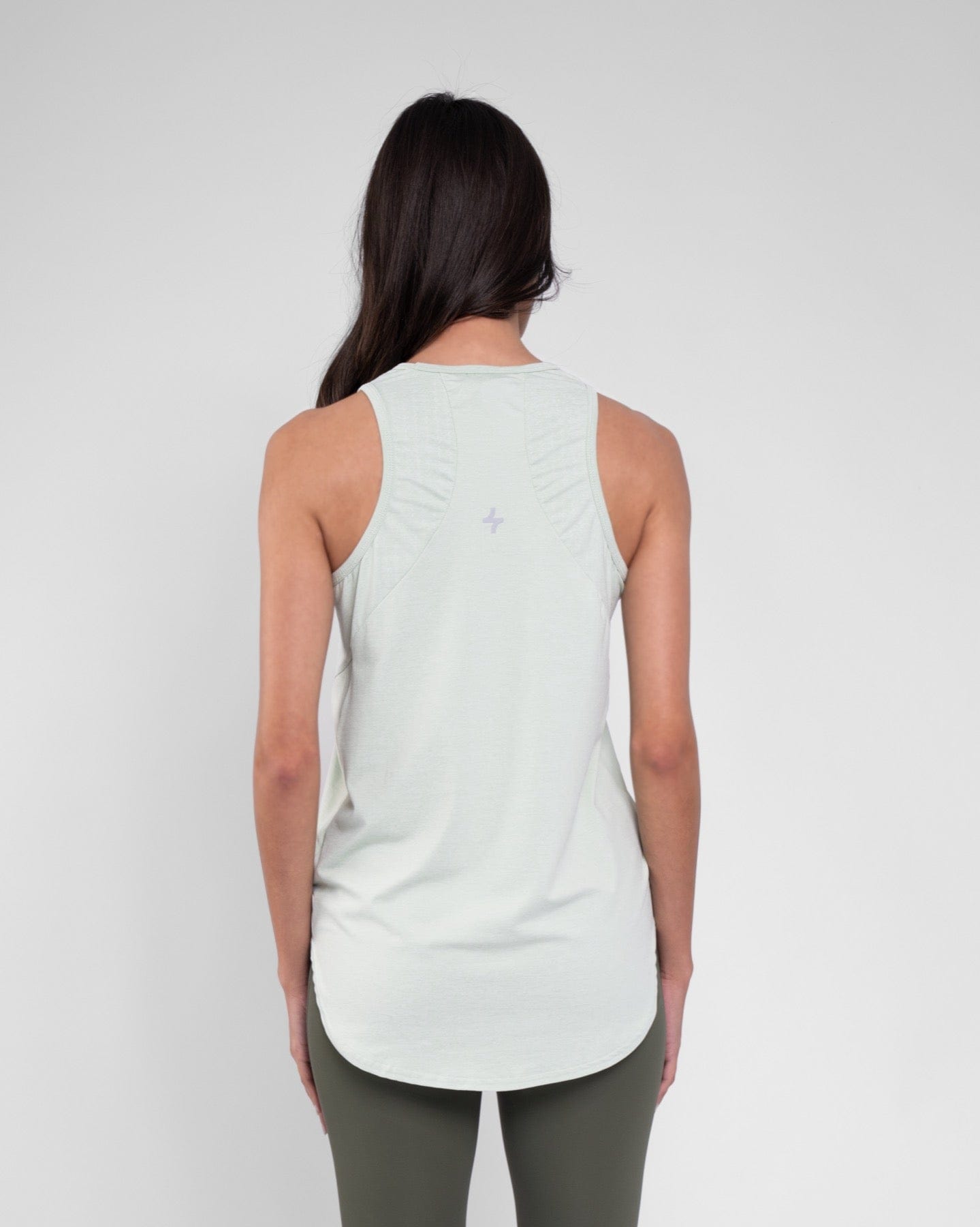 A woman standing with her back turned towards the camera, wearing a Sage modest activewear by qynda, sleeveless RUH TANK TOP and leggings against a neutral gray background