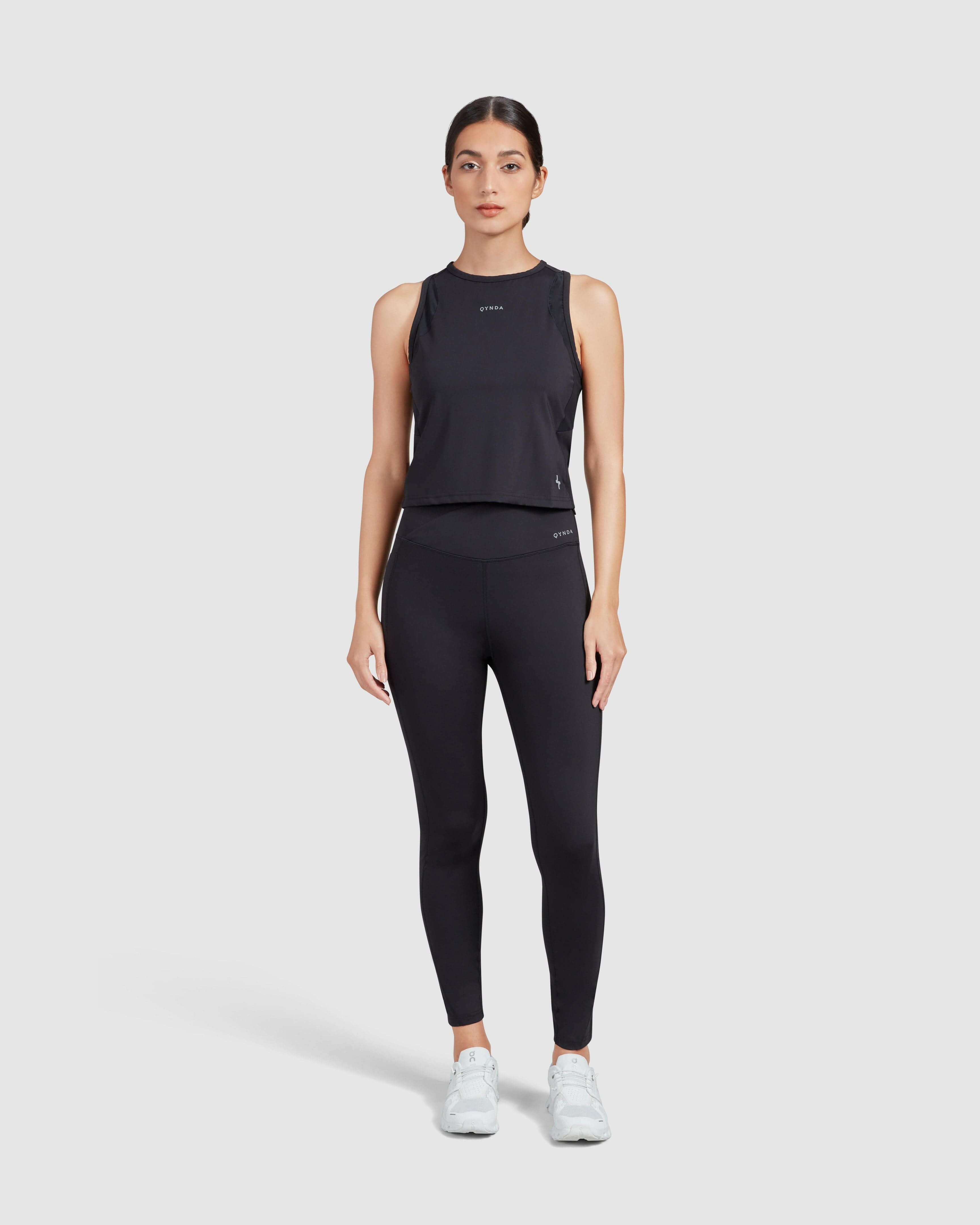 A woman standing confidently, wearing a qynda sporty black low impact gym tank top with a high neckline and leggings, paired with white sneakers, on a plain white background. Ideal for showcasing casual athletic wear.