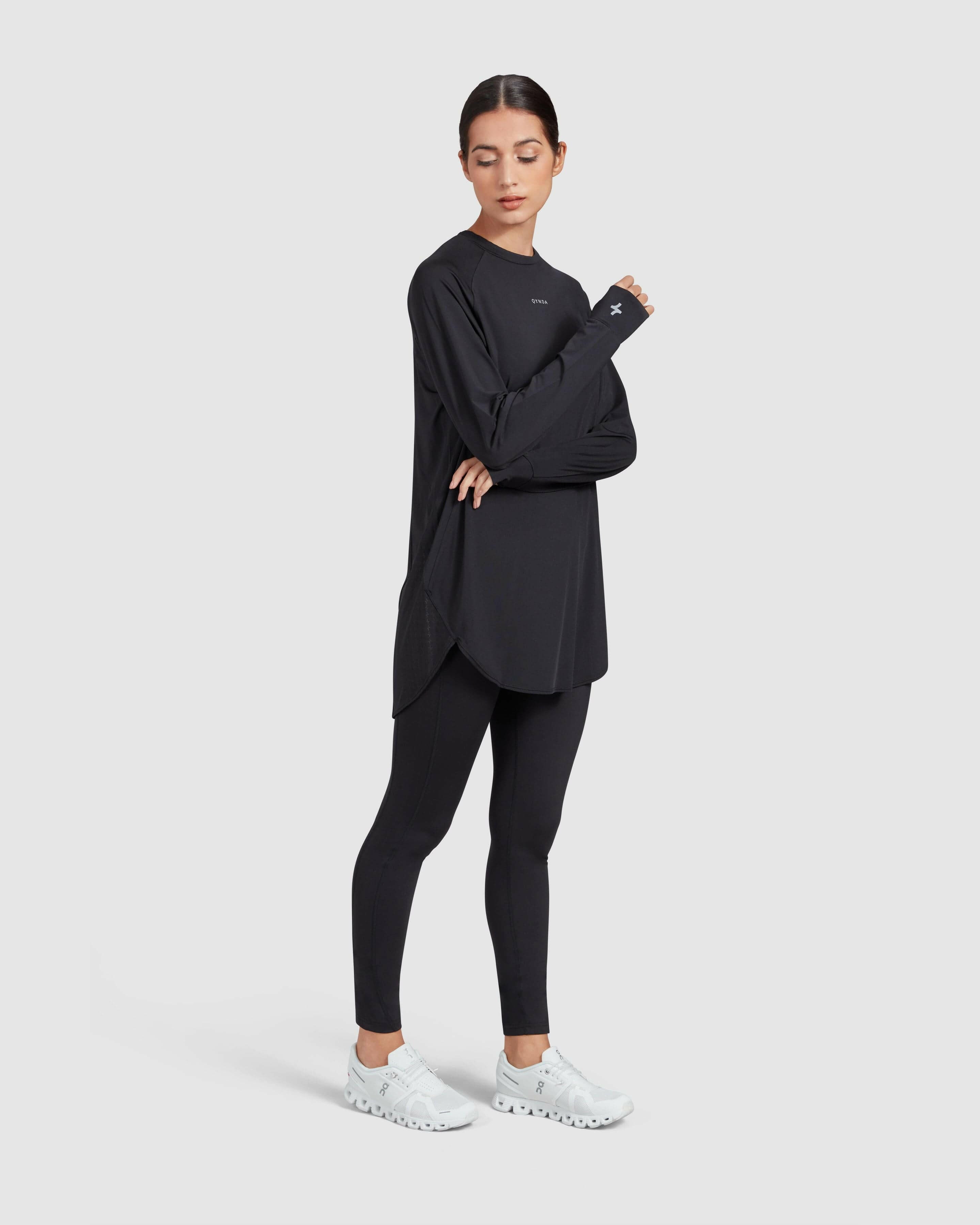 A model stands confidently in a modest Black MOD LONG SLEEVE T-SHIRT by Qynda made of super light material, black leggings, and white sneakers, arms crossed, looking to her left on a plain background.