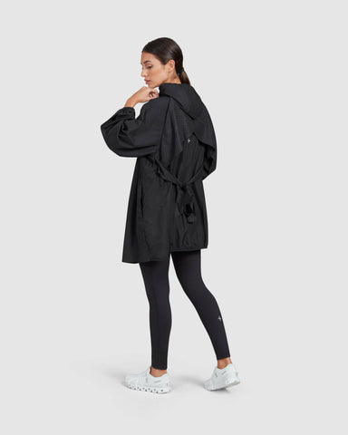 Woman in a casual pose wearing a black COOLDOWN coat by Qynda and leggings with white sneakers, looking away thoughtfully.