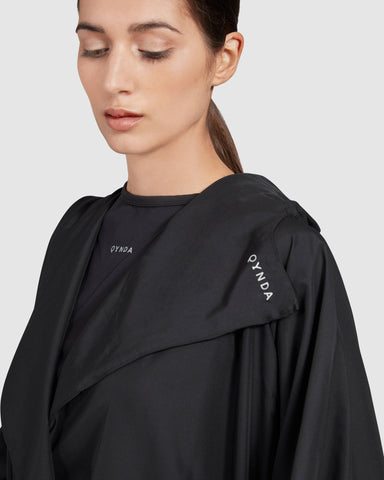 A woman in a black COOLDOWN coat COOLDOWN COAT with with Qynda brand logo on the shoulder, looking down thoughtfully.