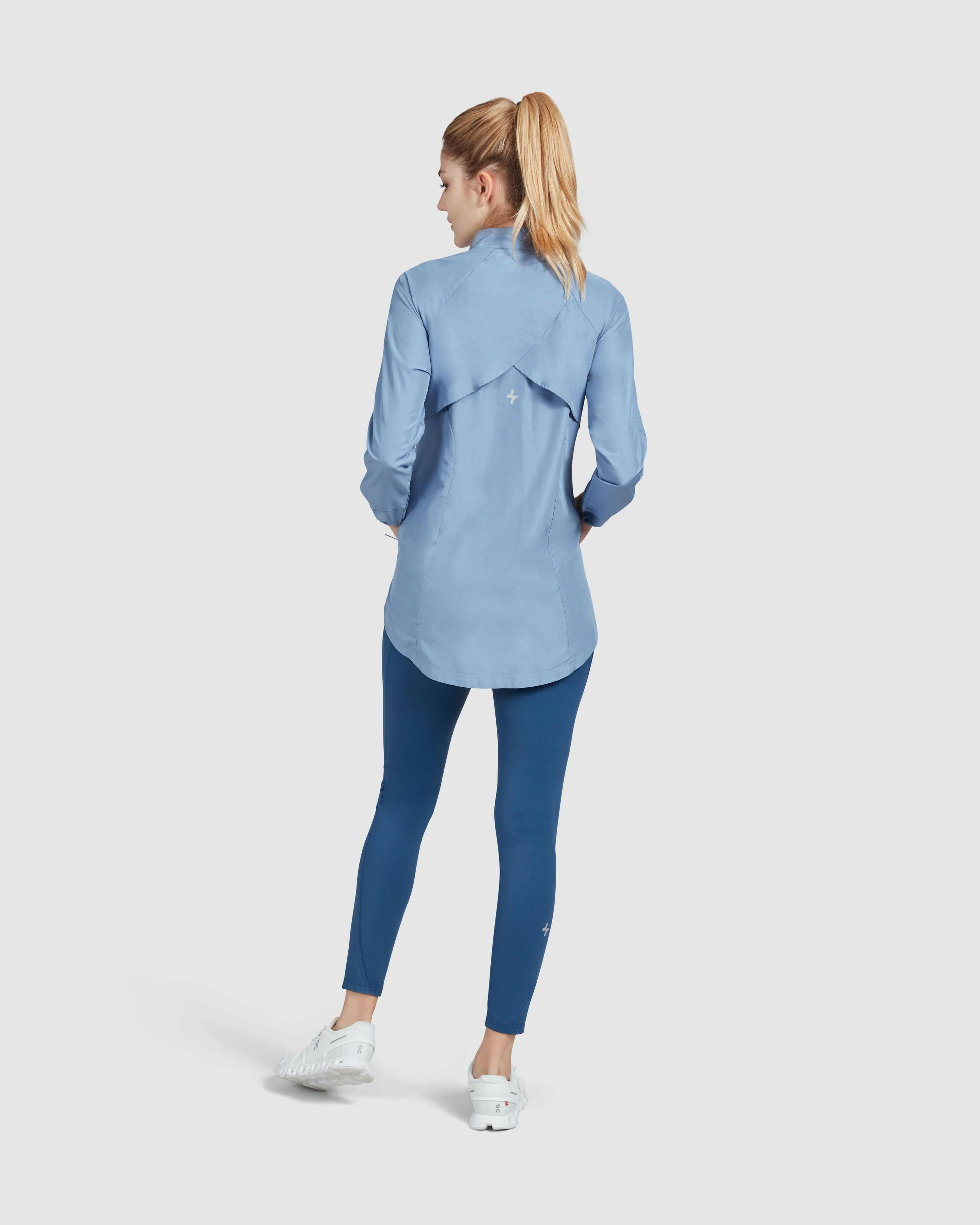A model seen from behind, standing against a neutral background, wearing a Infinity Blue FLY JACKET by Qynda with a hidden mesh back.