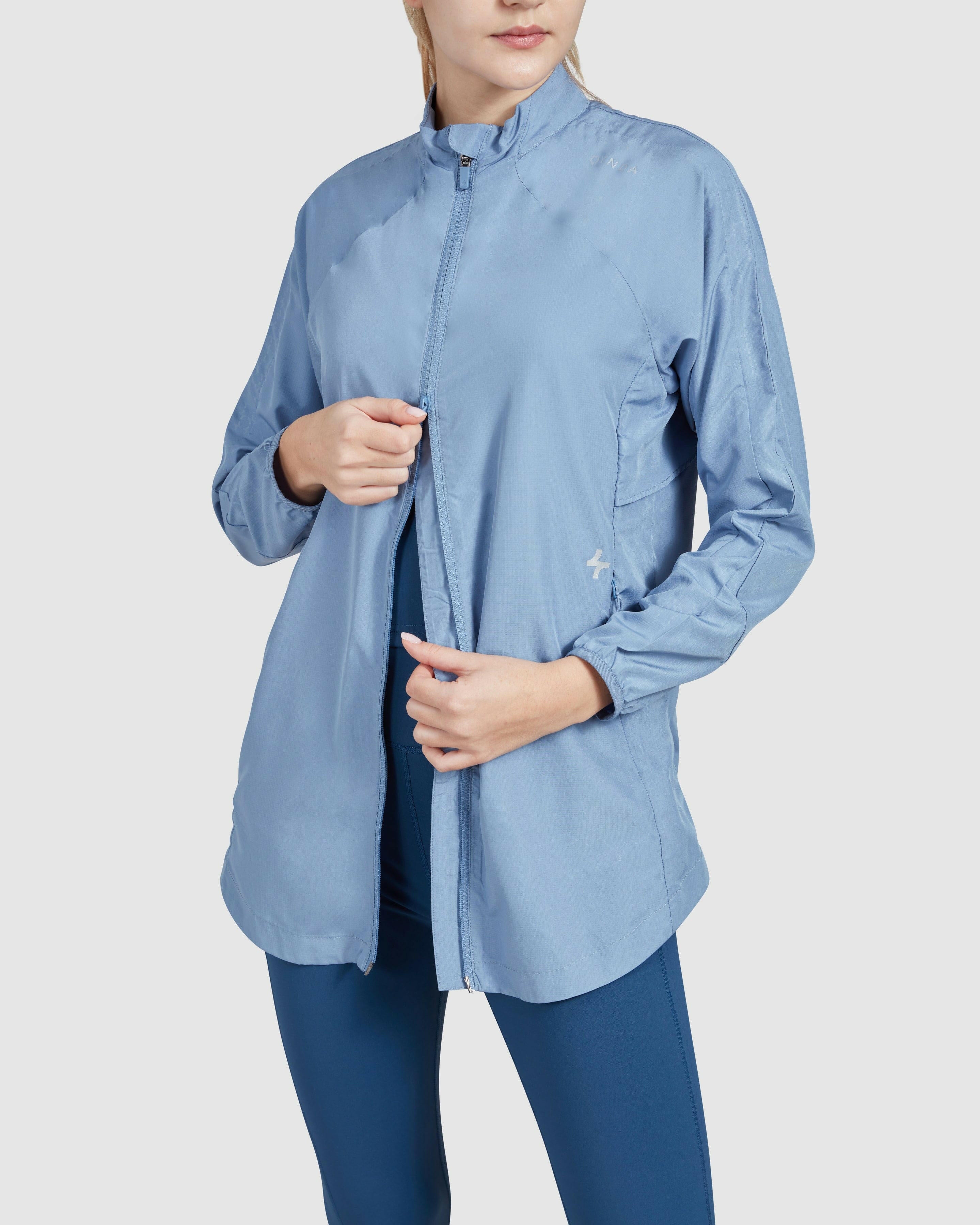 A woman is modeling an Infinity Blue, athletic zip-up FLY JACKET with a high collar, showcasing its style and fit.