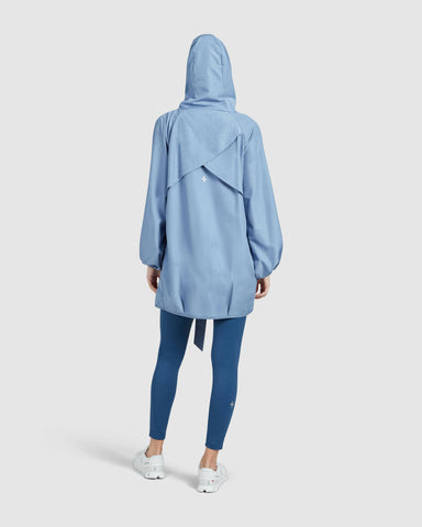 Woman standing with their back to the camera, wearing a hooded Infinity Blue COOLDOWN coat by Qynda over leggings and white sneakers.