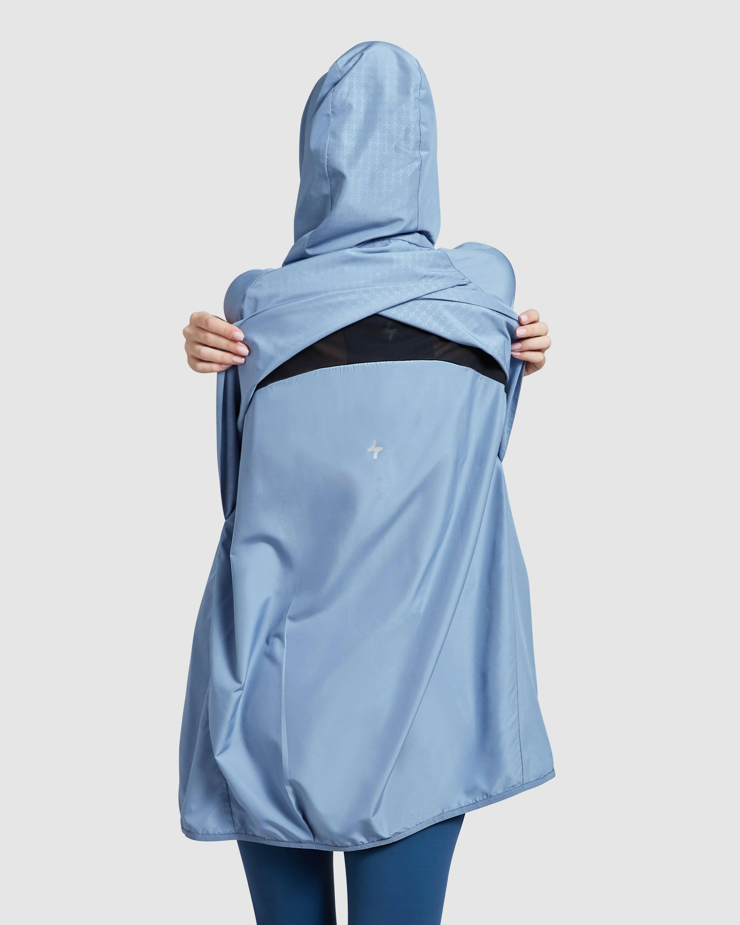 A woman seen from behind wearing a COOLDOWN coat by Qynda in Infinity Blue, made of super light fabric, with a hood casually slung over the shoulder to reveal a black inner lining, against a plain