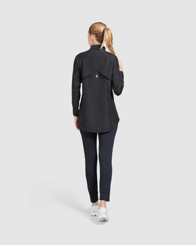 Back view of a model standing poised in a casual black long-sleeve FLY JACKET by Qynda and dark leggings paired with white sneakers, against a plain light background.