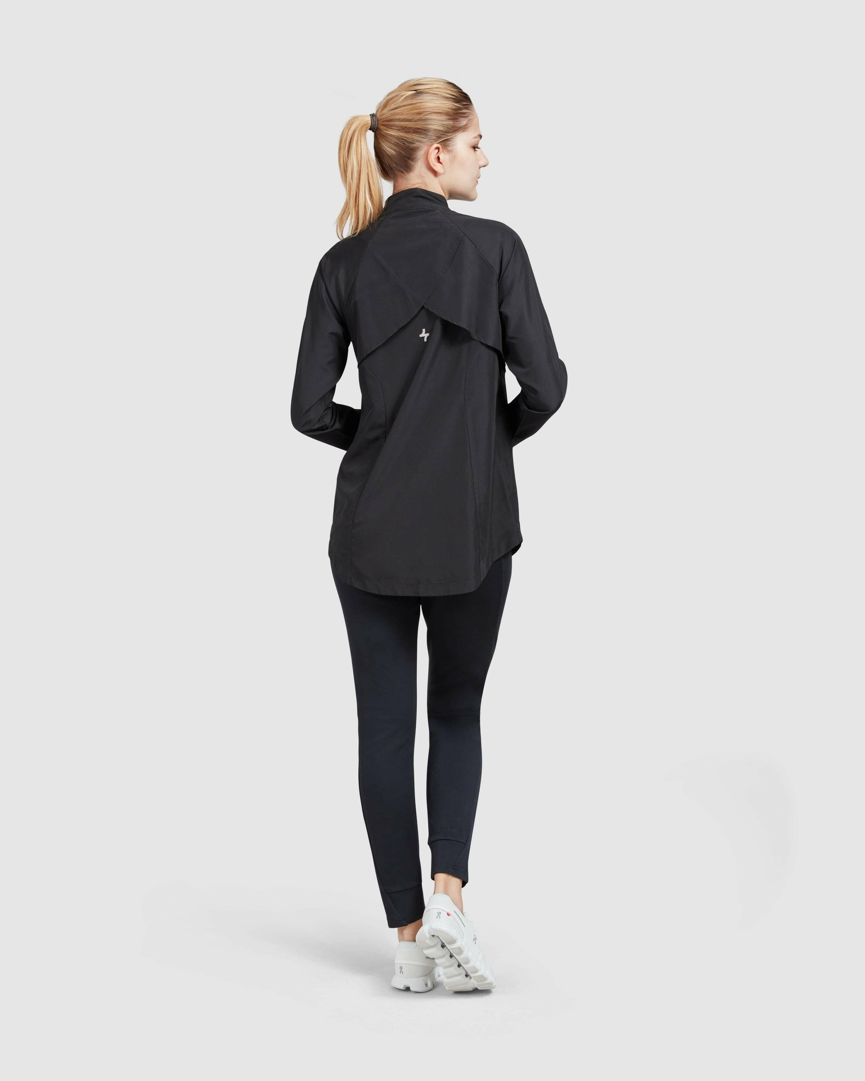 Back view of a model standing poised in a casual black long-sleeve FLY JACKET by Qynda and dark leggings paired with white sneakers, against a plain light background.