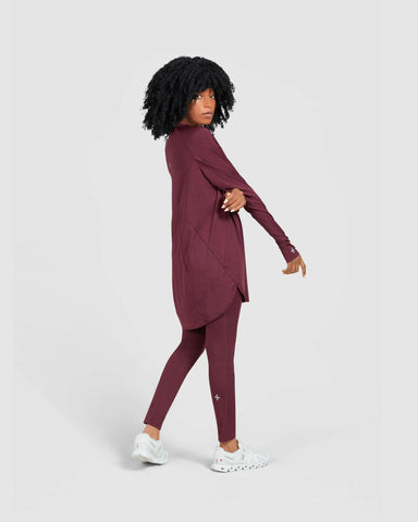 Young woman in a modest maroon MOD LONG SLEEVE T-SHIRT by Qynda made of light material and white sneakers, walking to the left and looking over her shoulder against a white background.