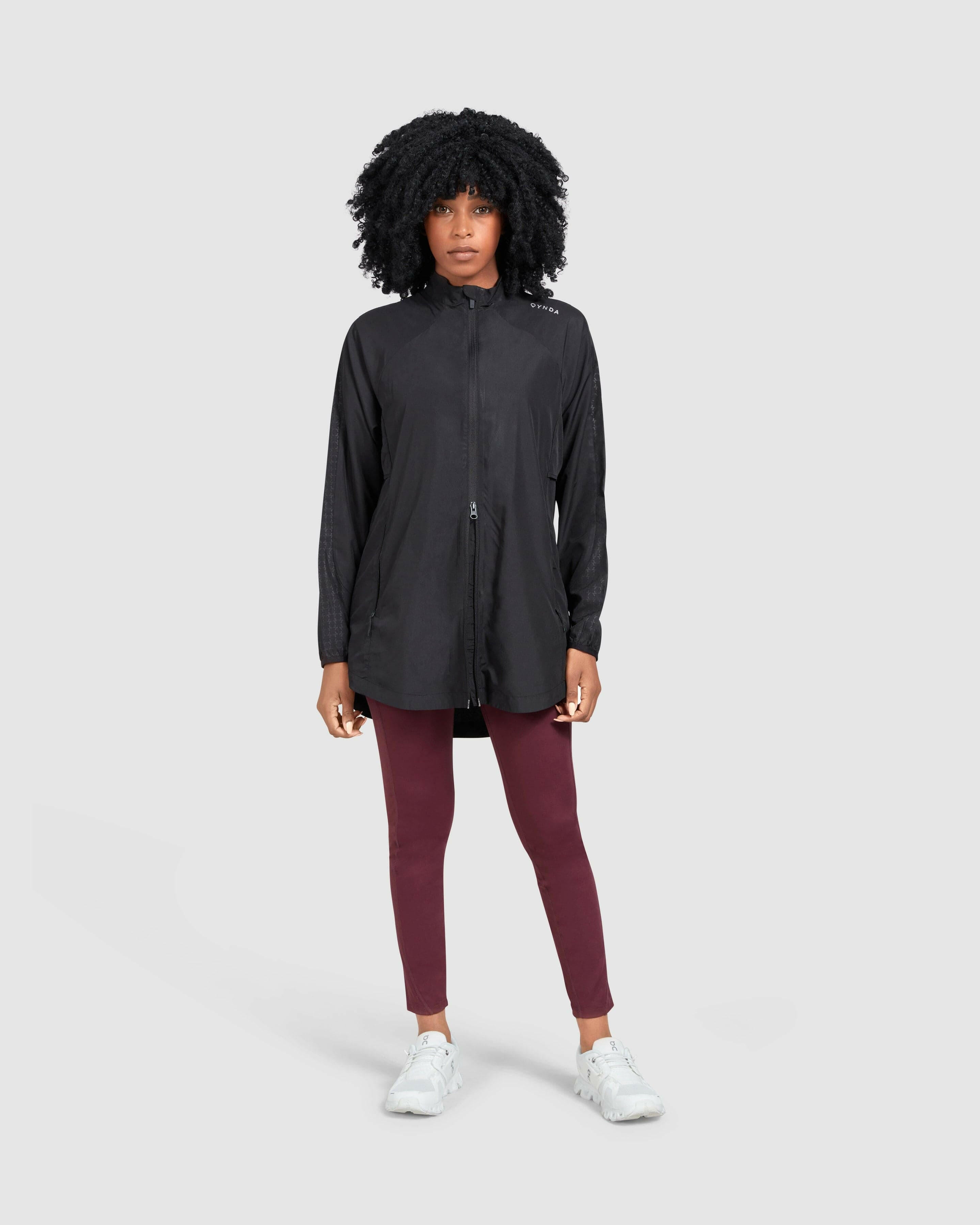 A confident model in a black FLY JACKET by Qynda made of super light fabric and maroon leggings designed for a bigger motion range, paired with casual white sneakers, standing against a neutral background with a focused gaze.