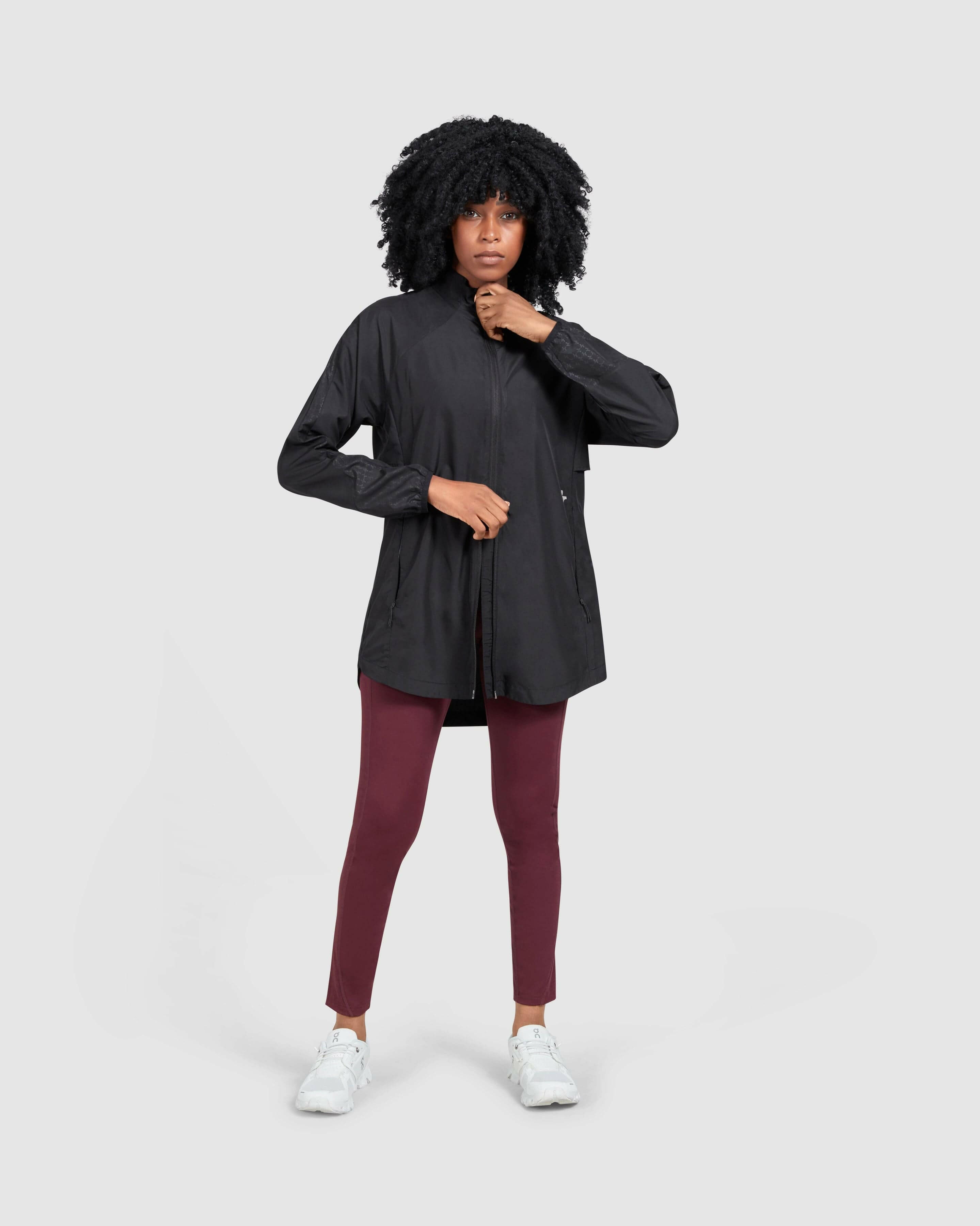 A confident model in a black FLY JACKET by Qynda made of super light fabric with a hidden mesh back and both ways zipper and maroon leggings.