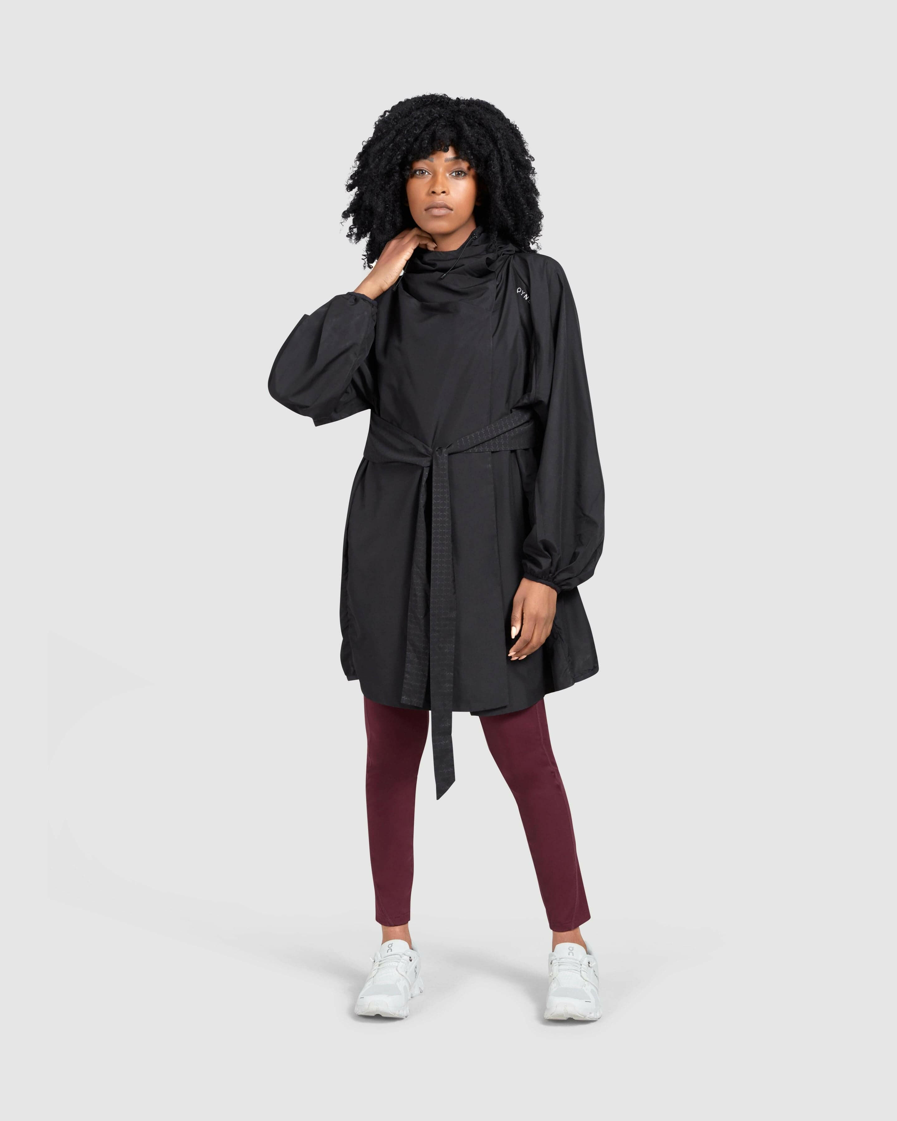 A woman in a stylish black COOLDOWN coat by Qynda, maroon leggings, and white sneakers, posing confidently against a white background.