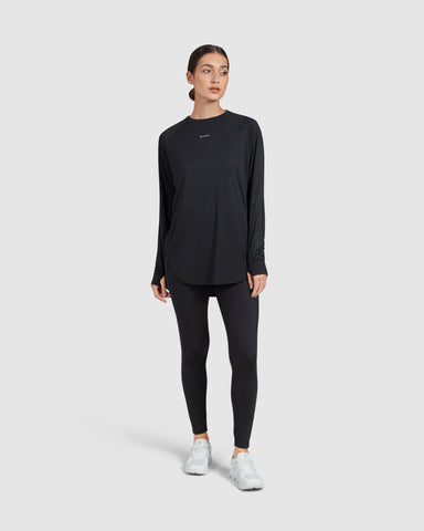 A woman standing against a plain background, dressed in an active long-sleeve t-shirt by QYNDA, black leggings, and white sneakers, epitomizing a minimalist athletic style with no see