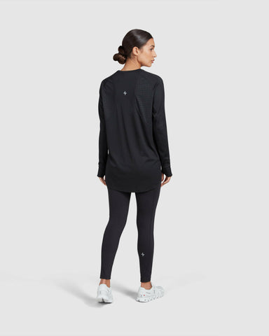 A woman stands turned to the side against a plain background, dressed in an act long-sleeve t-shirt by QYNDA, black leggings, and white sneakers.