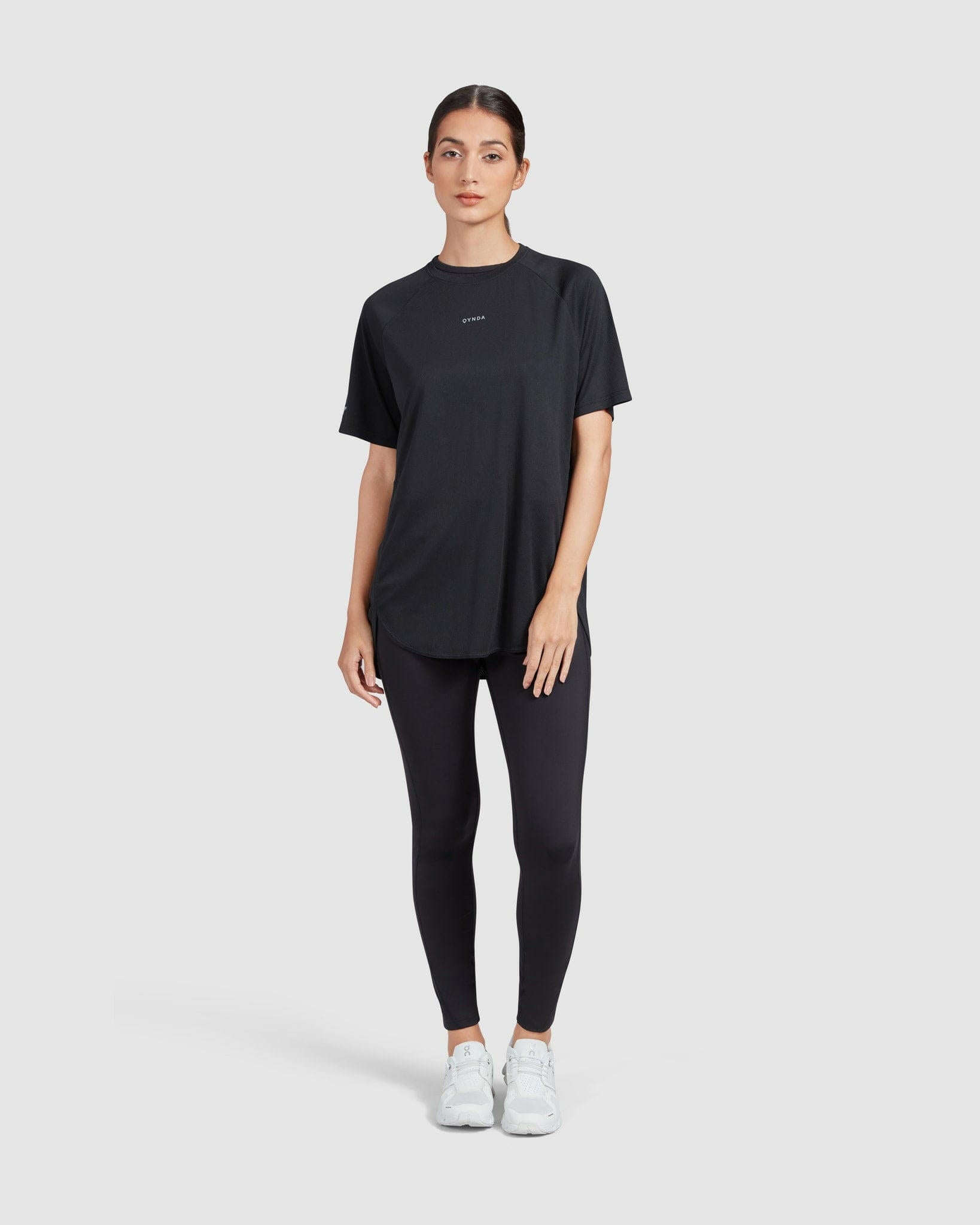 A woman stands against a white background, wearing a casual black short sleeve BREATHE T-SHIRT by Qynda, black leggings, and white sneakers designed for breathability and running, looking straight ahead with a neutral expression.