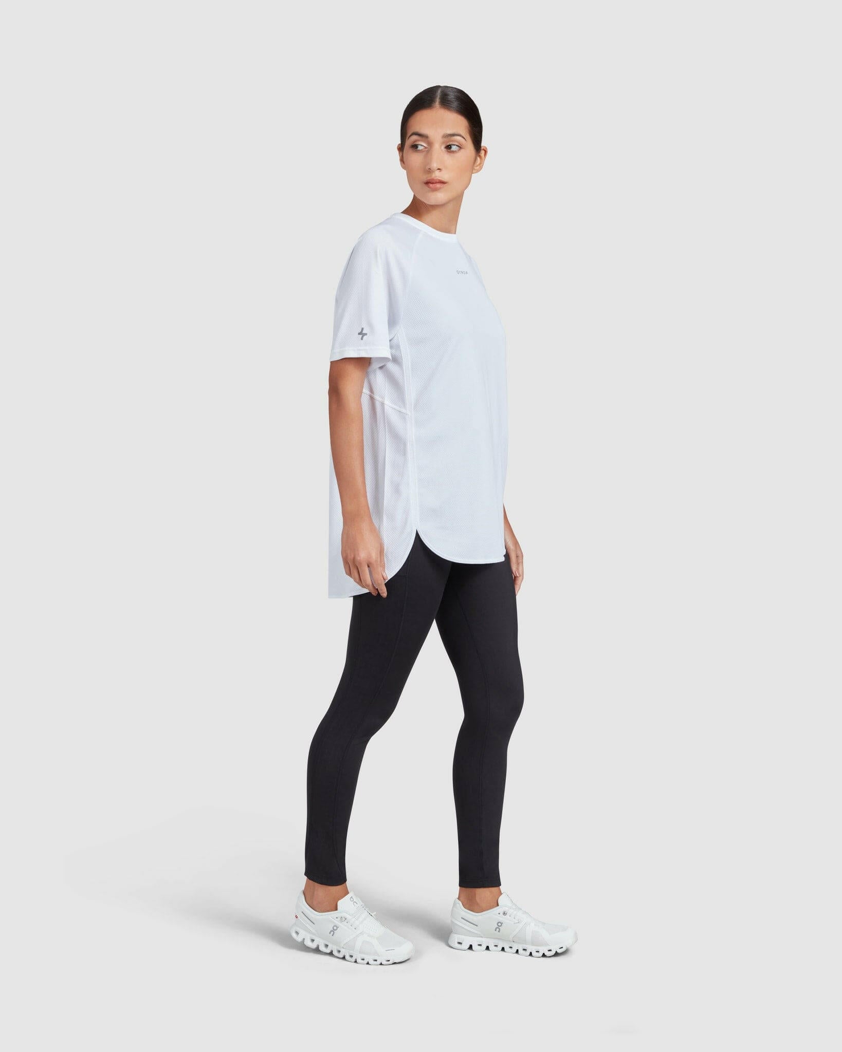 A woman in a white short sleeve t-shirt and black leggings standing against a white background, giving a side pose while gazing into the distance, exemplifying a casual, sporty, and breathable look