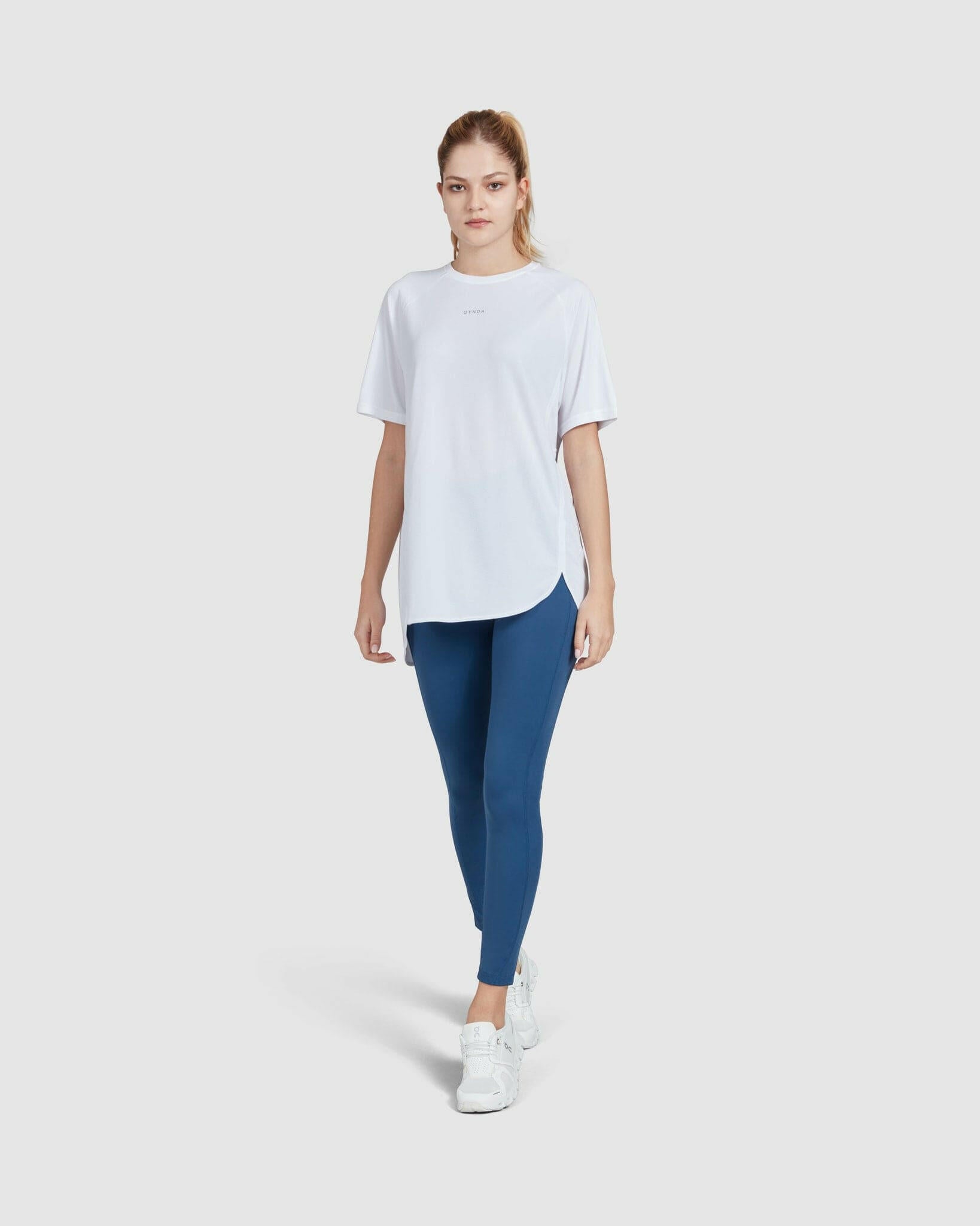 A young woman in a casual short sleeve white BREATHE T-SHIRT by qynda and blue leggings standing against a white background, wearing white sneakers, embodying a simple, sporty, and comfortable style.