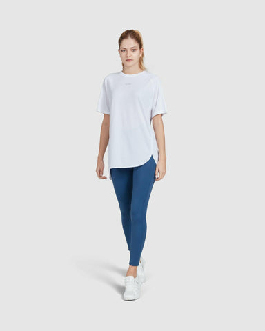 A young woman in a casual short sleeve white BREATHE T-SHIRT by qynda and blue leggings standing against a white background, wearing white sneakers, embodying a simple, sporty, and comfortable style.