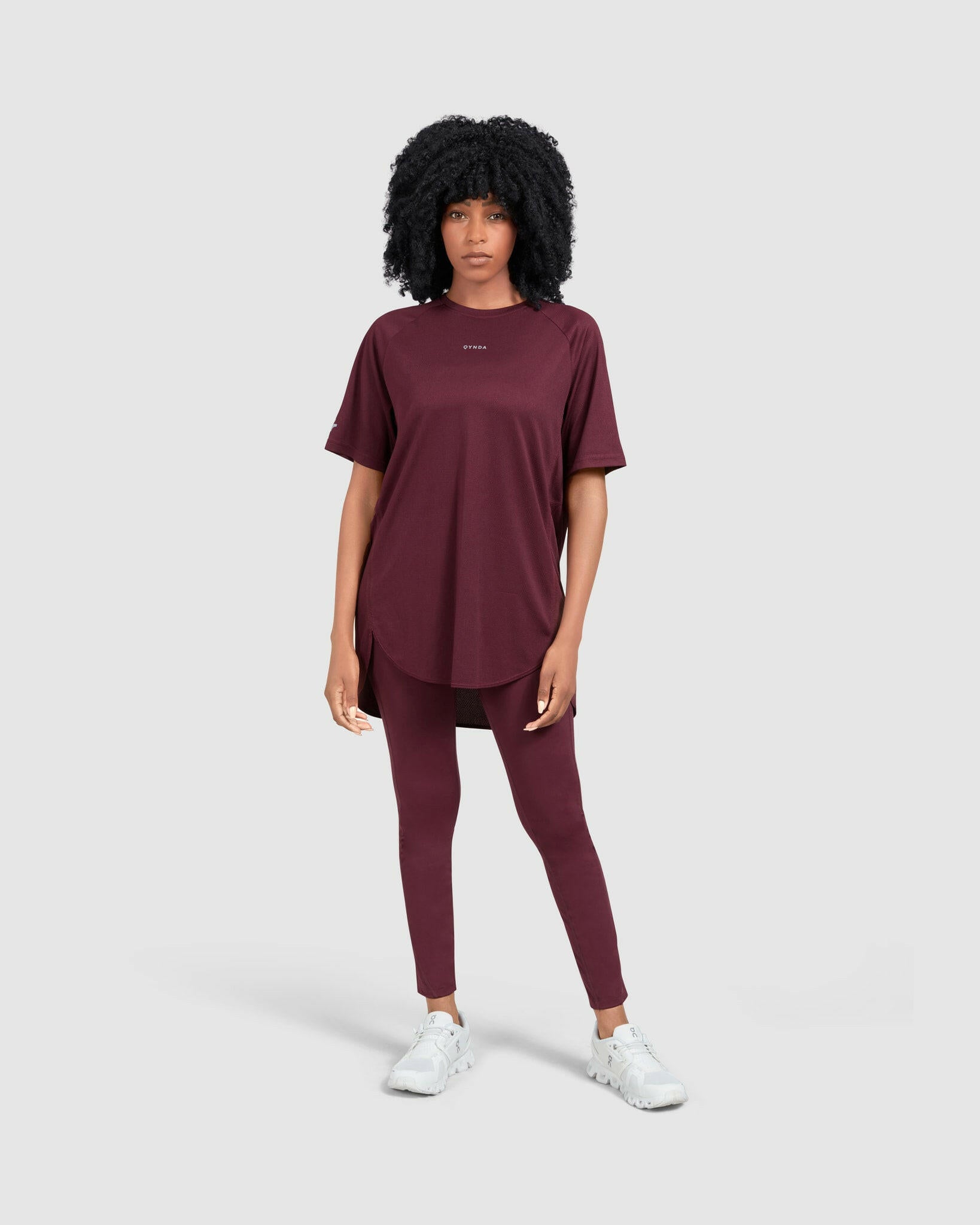 A woman stands against a neutral background, modeling a casual maroon activewear outfit comprising a short sleeve BREATHE T-SHIRT, by Qynda brand, designed for breathability, and leggings, paired with white sneakers. Her hair is styled
